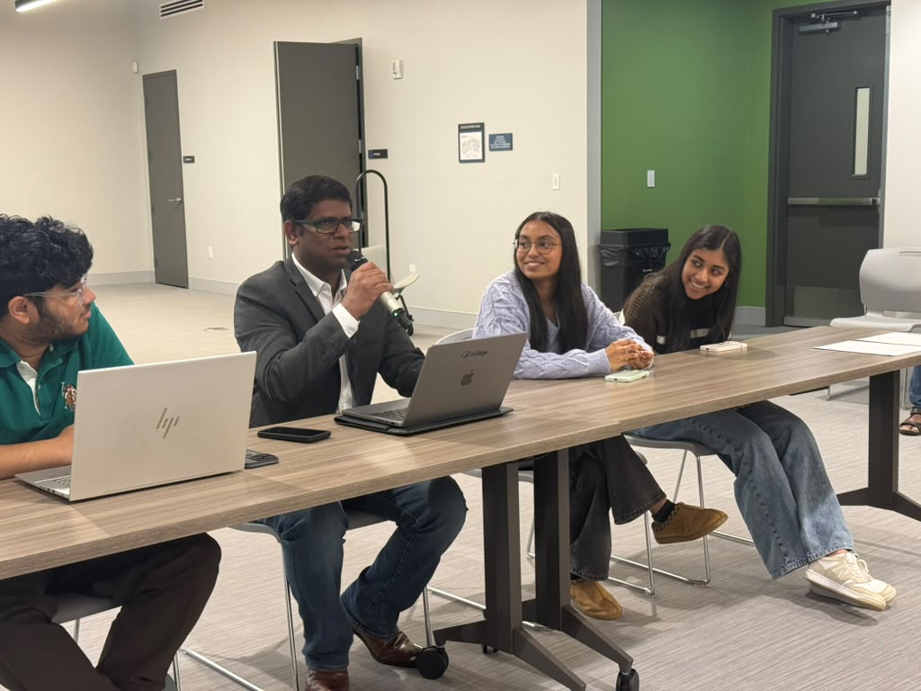 Four people sitting at a long table in a meeting room, with laptops and notebooks, engaged in discussion.