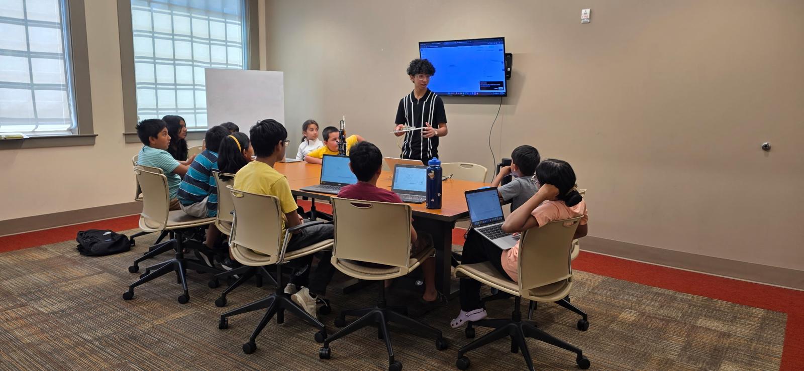 A classroom with students seated around a table, a teacher standing at the front, and a screen on the wall.