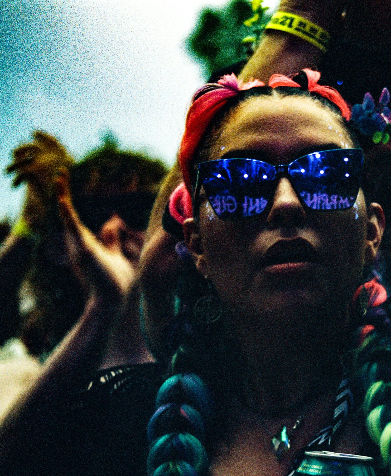 Person wearing sunglasses with reflections, colorful braided hair, and a headband, in a crowd outdoors.
