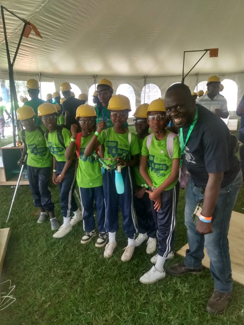 Group of children wearing yellow helmets and green shirts, standing with an adult inside a tent, smiling.