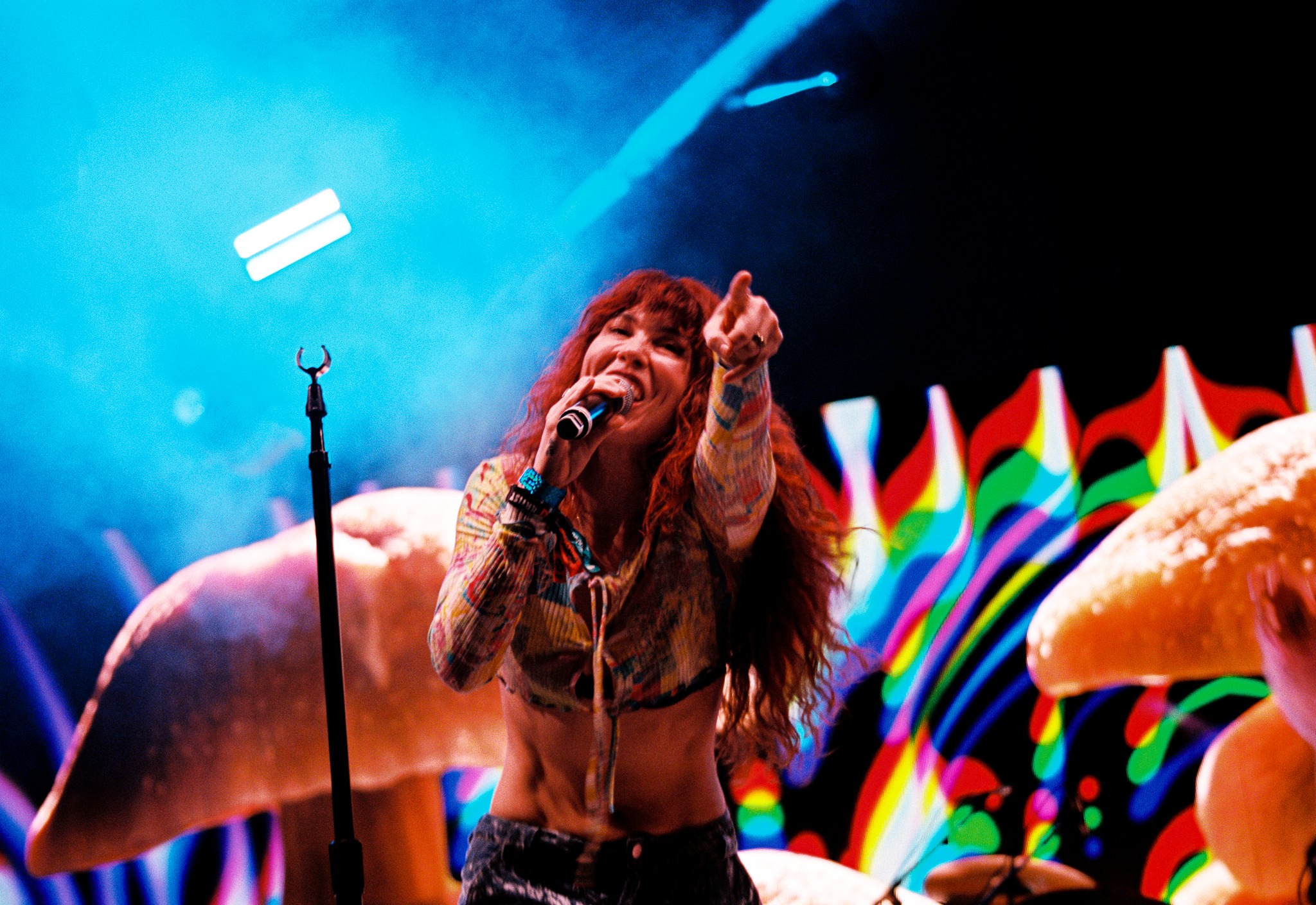 Performer with long hair singing into a microphone on stage with rainbow flags and colorful background.