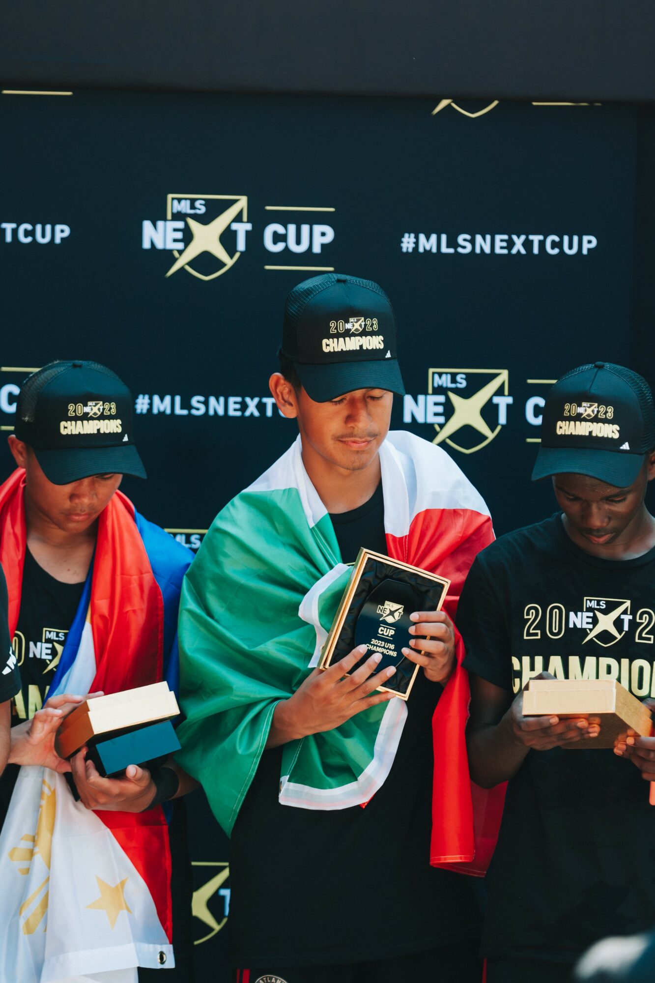 Three young men wearing caps and holding awards, standing in front of a backdrop with 'MLS NEXT CUP' and 'CHAMPION' text.