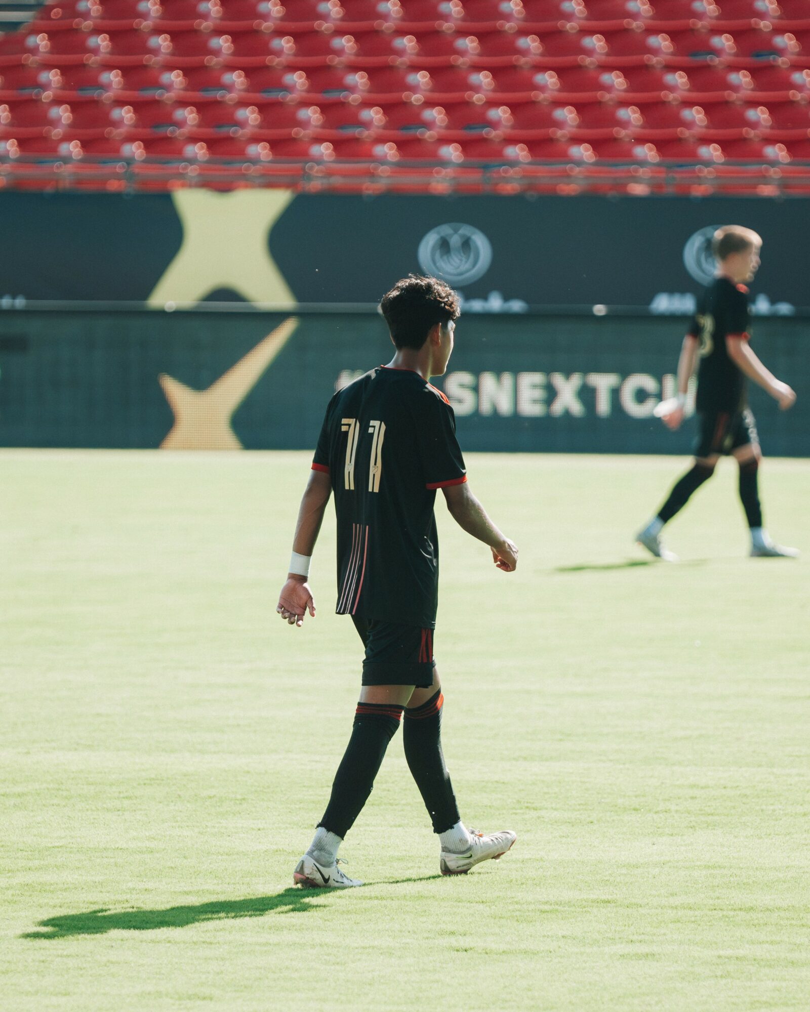 Child in black sports uniform walking on a soccer field with other players in the background.