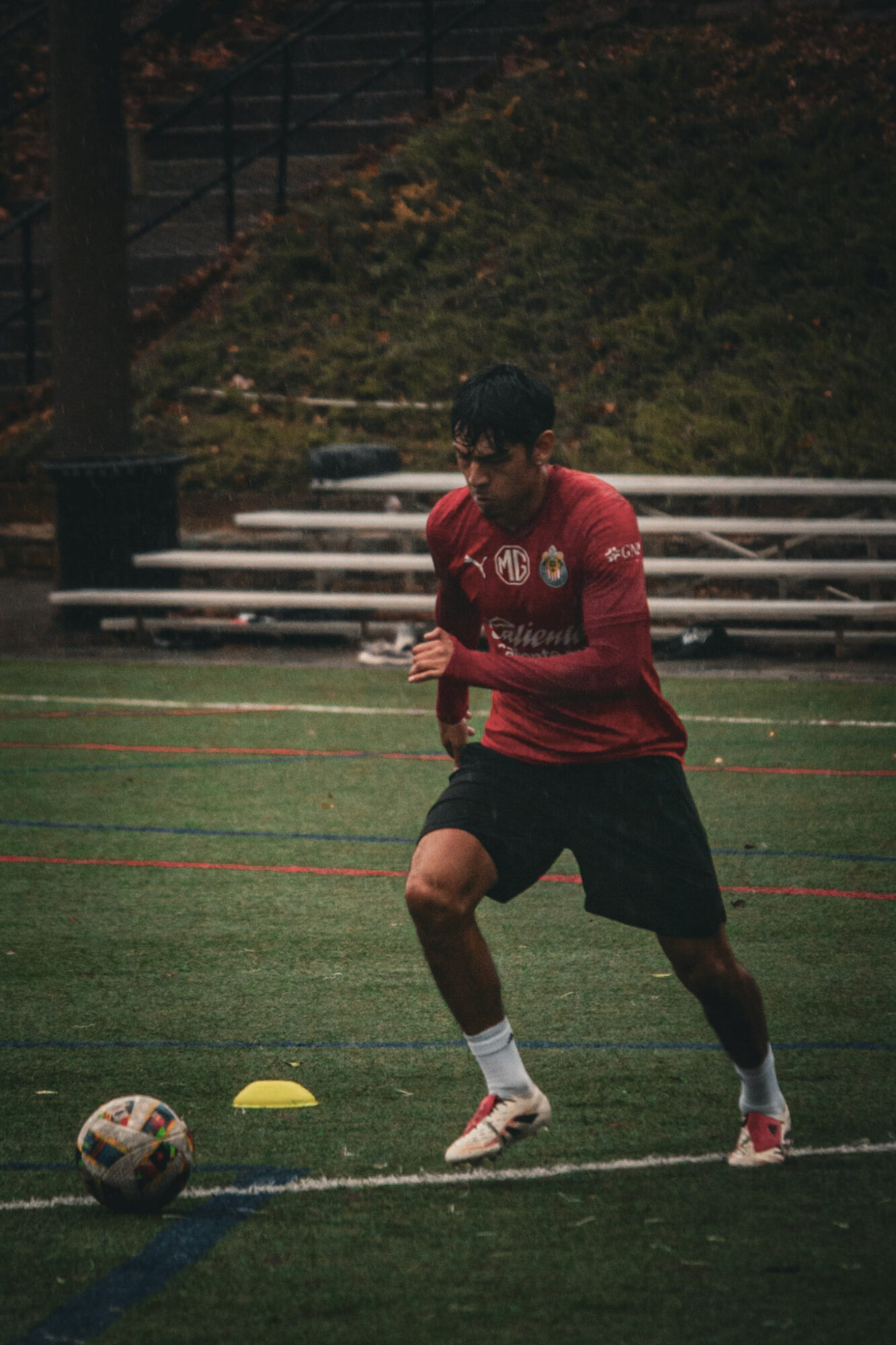 Person in red shirt and black shorts playing soccer on a field with trees in the background.