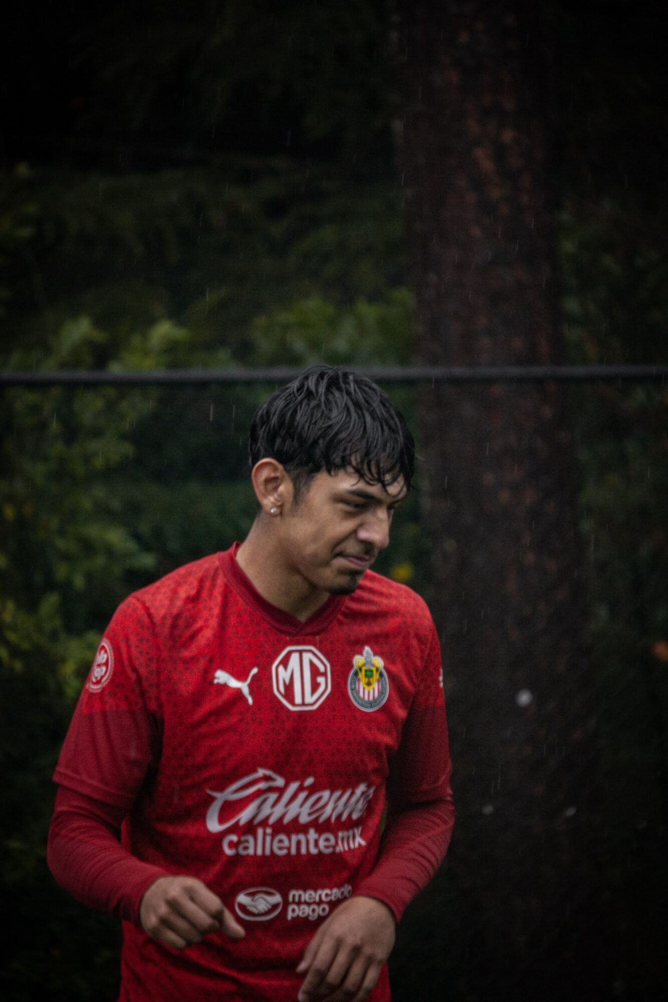 Young man in red sports jersey standing outdoors with trees and a fence in background.