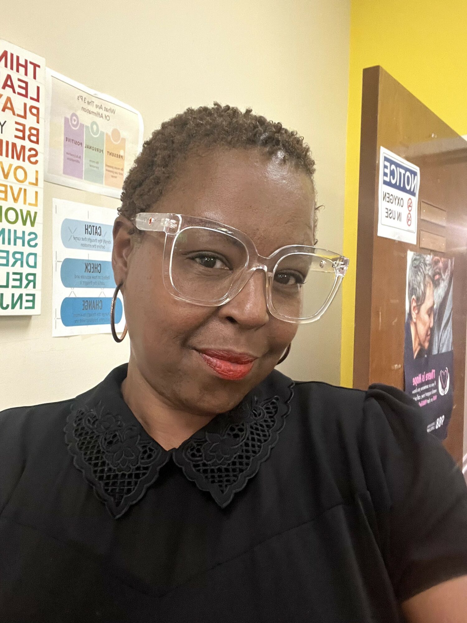 Woman with short curly hair wearing glasses and a black top, standing indoors near posters and a yellow wall.
