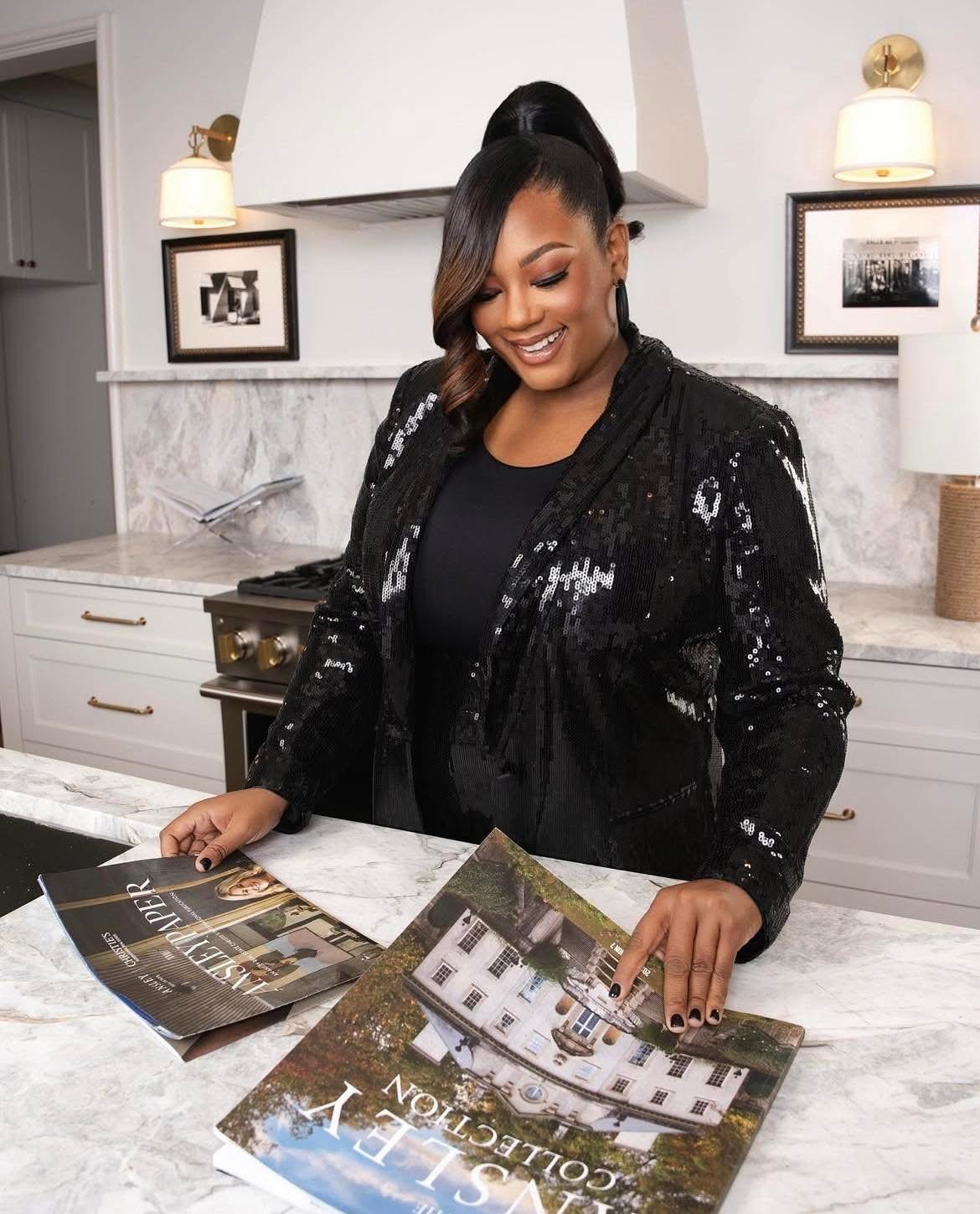 Smiling woman in black sequined jacket looking at magazines on kitchen counter, with modern white cabinets and framed photos in background.