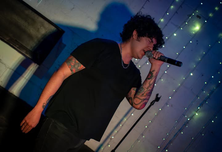 Young man with curly hair singing into a microphone, wearing a black shirt, tattoos visible on arms, stage with blue lights.
