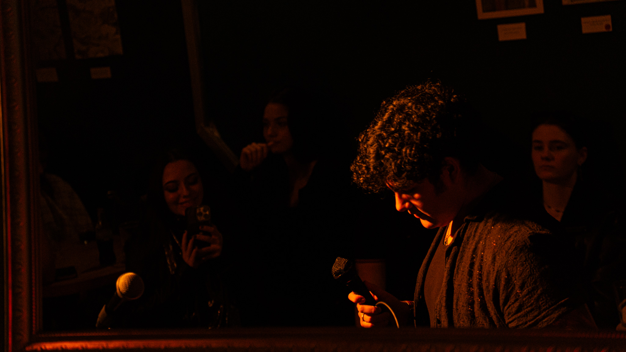 Person with curly hair looking at a phone in a dark setting, with a group of people in the background.