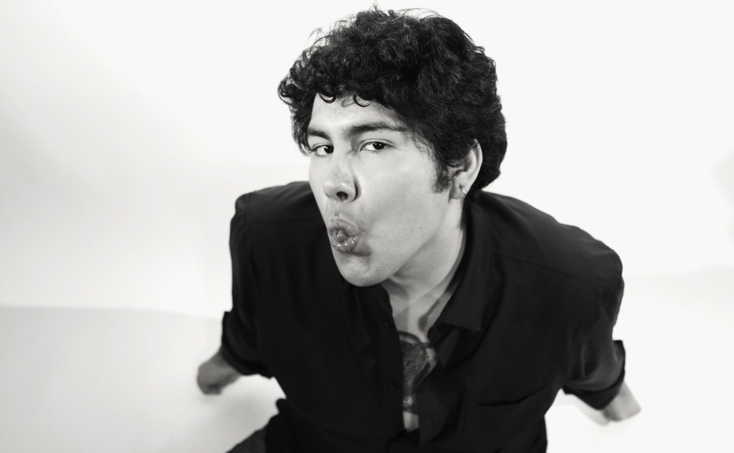 Young man with curly hair making a surprised face, leaning forward, wearing a dark jacket, against a plain background.