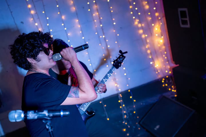 Person with curly hair singing into a microphone and holding an electric guitar, stage with string lights in background.