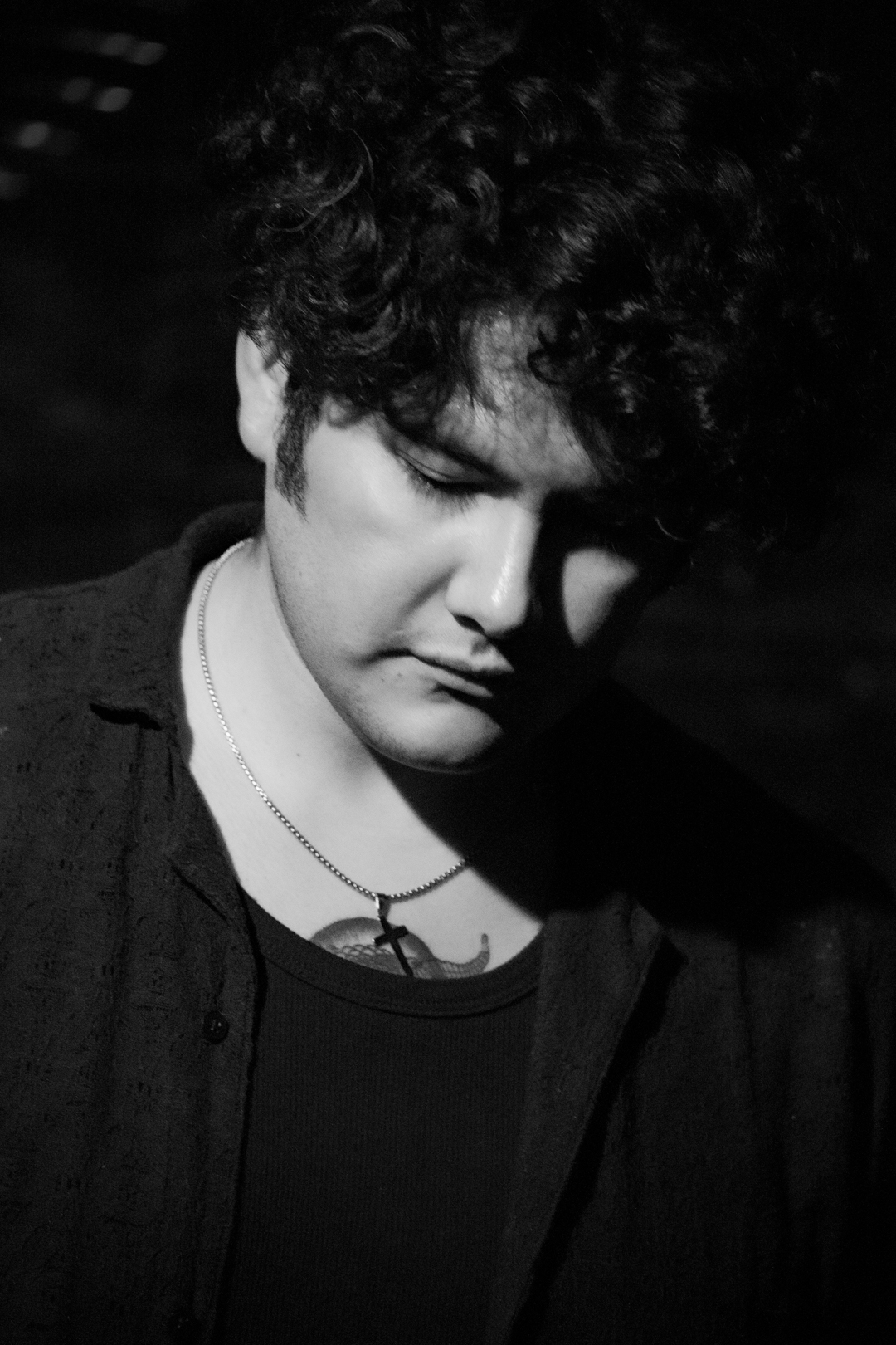 Young person with curly hair looking downward, wearing a necklace and dark clothing, in black and white.