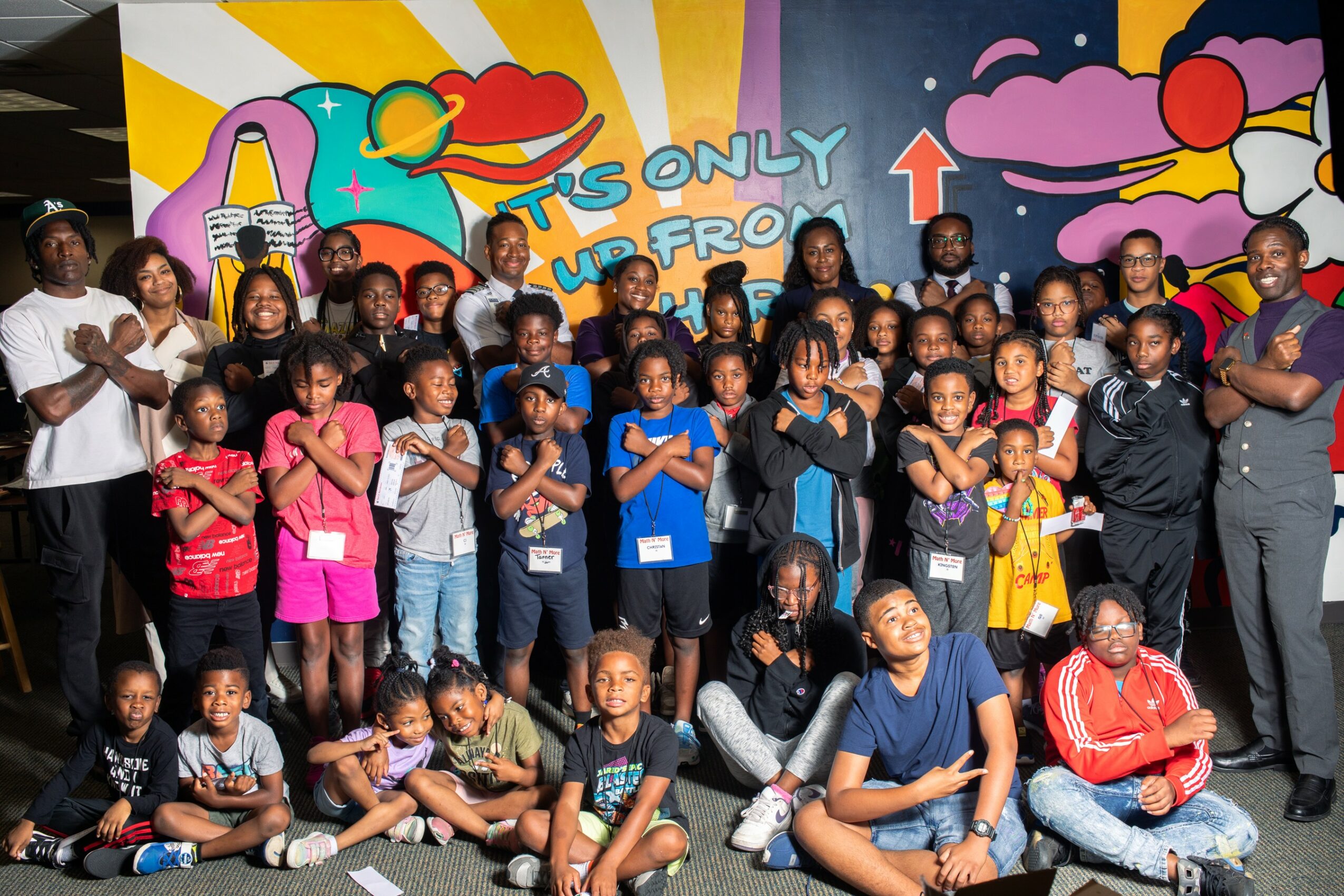 Group of children and adults posing in front of colorful mural with clouds, rainbow, and text, indoors.