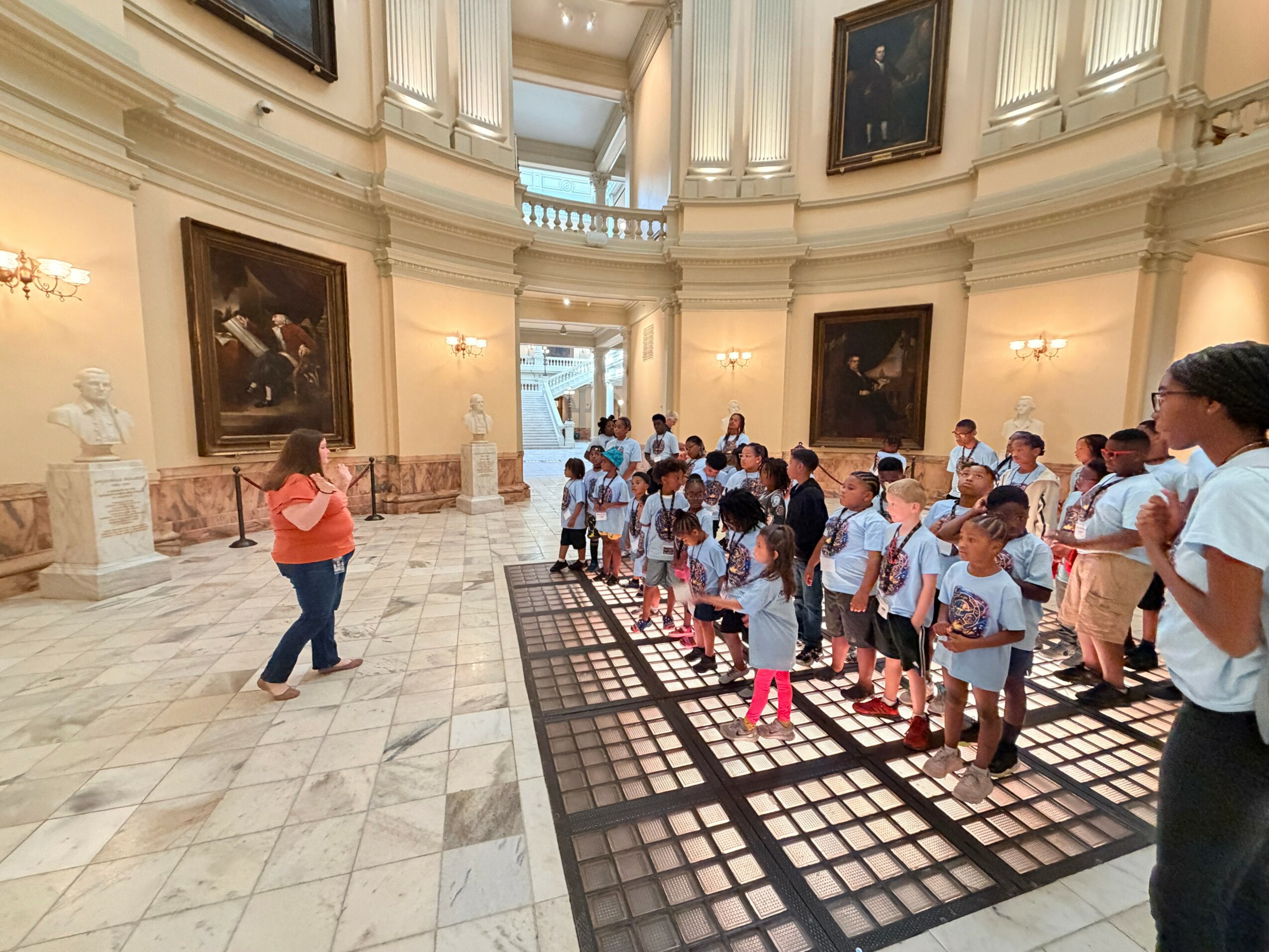 Group of children and adults inside a grand, ornate hall with paintings on the walls and marble floors.
