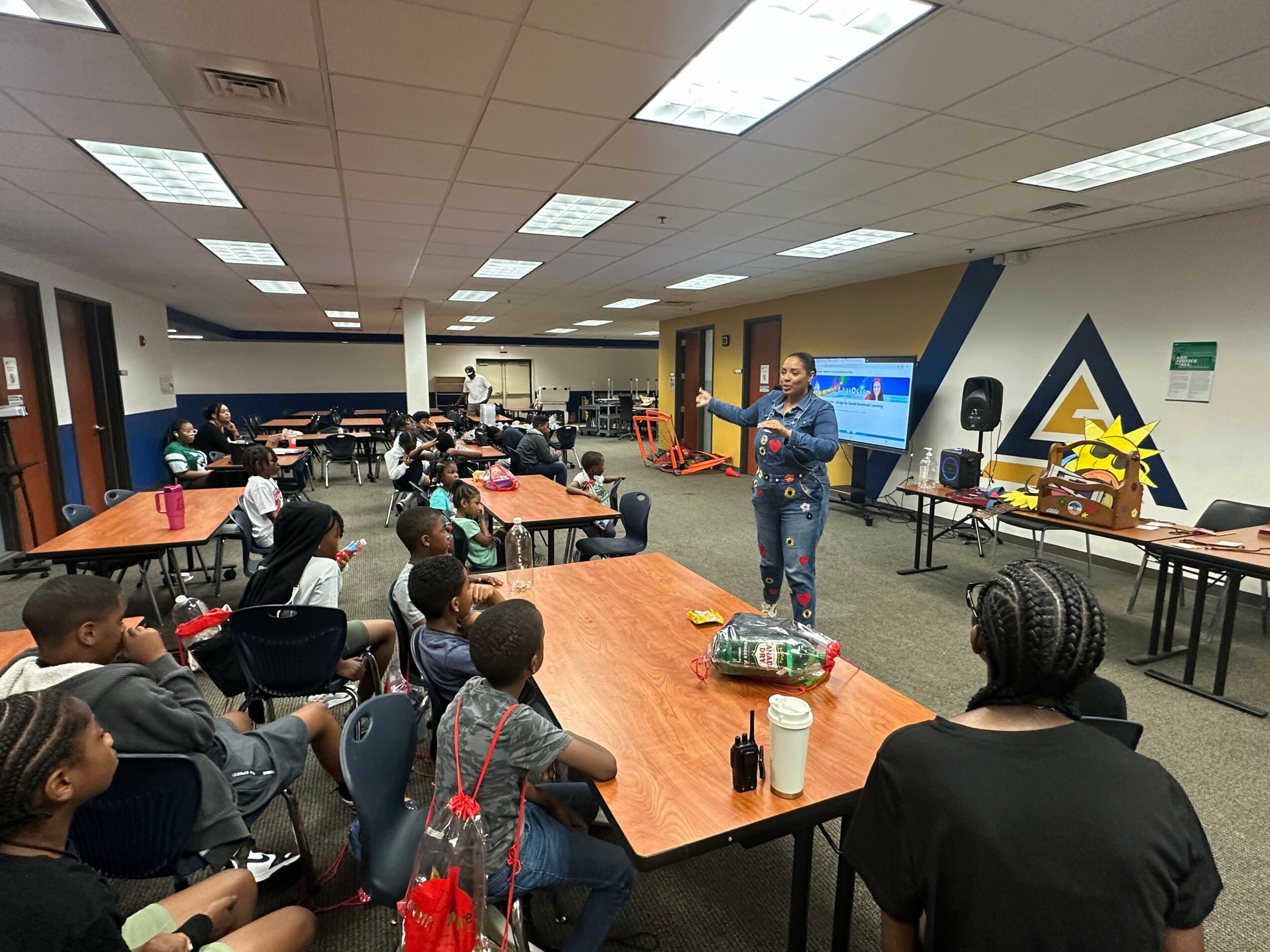 A person stands in front of a group of children and adults in a classroom or conference room, giving a presentation.