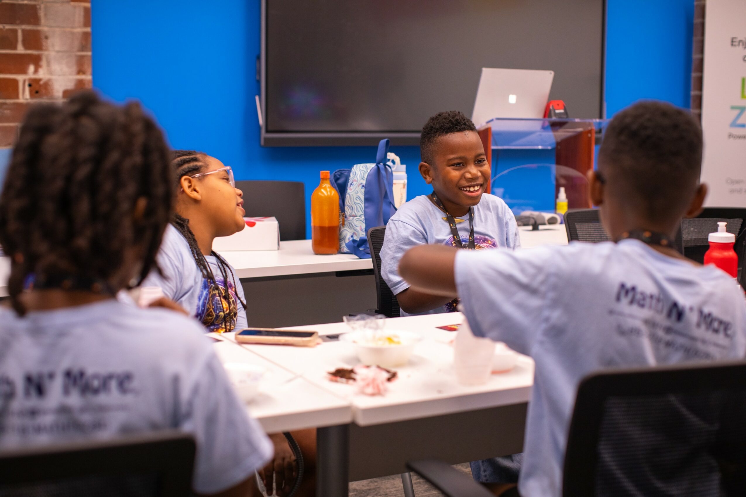 Group of children sitting at a table in a classroom, smiling and engaging with each other, with a large screen and supplies in the background.