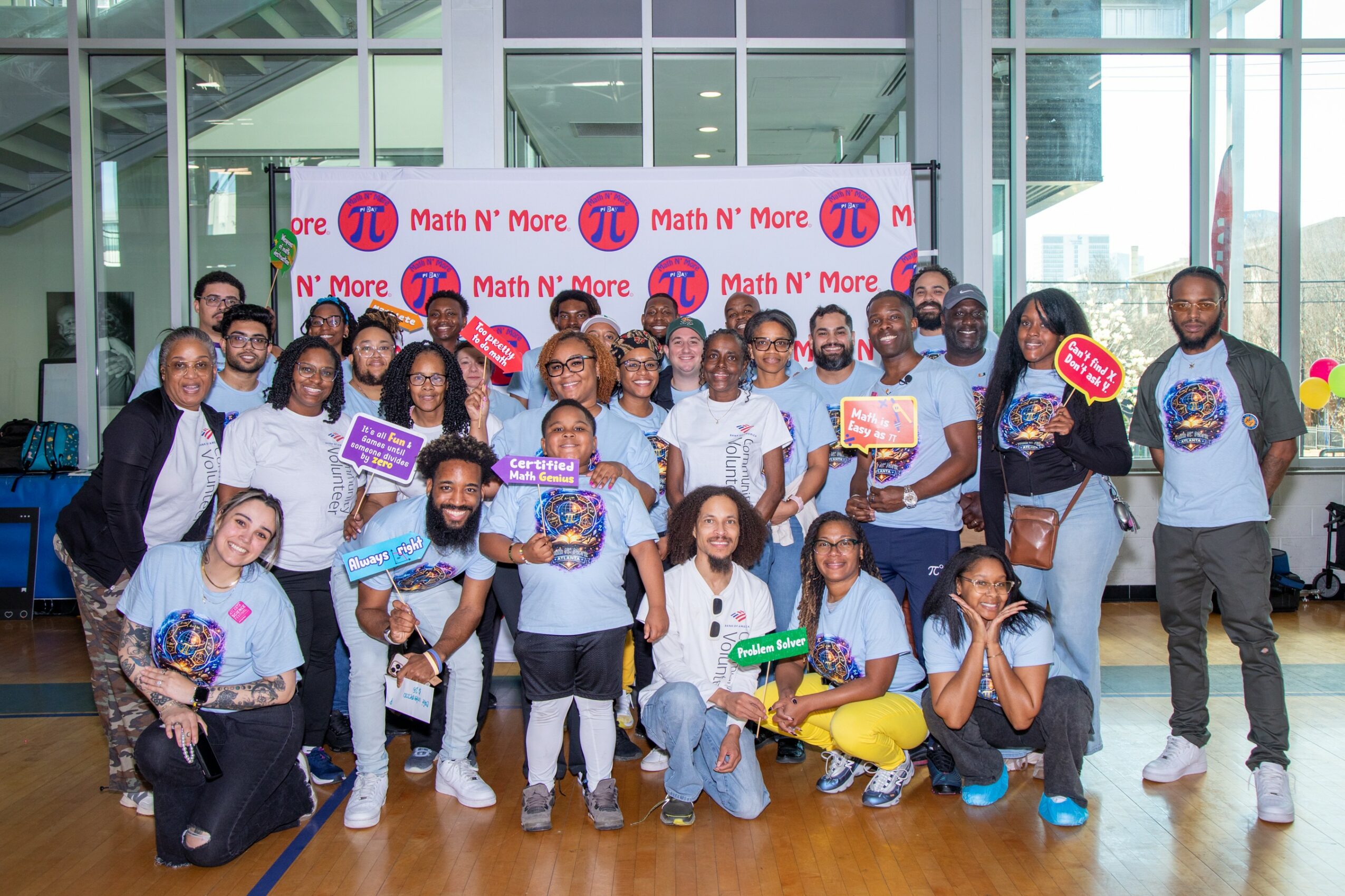 Group of children and adults posing indoors in front of a backdrop with 'Math N' More' logos, holding colorful signs and certificates.