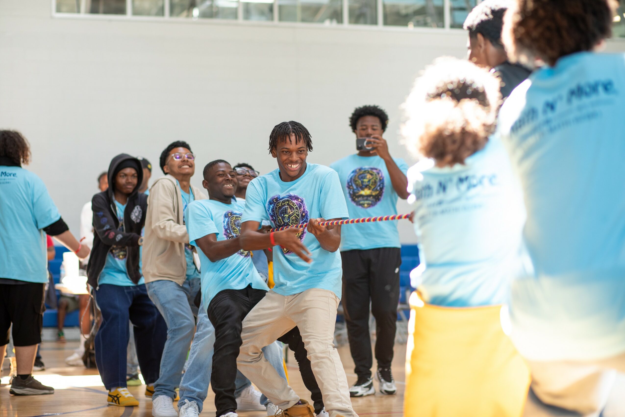 Group of young people playing tug-of-war outdoors, smiling and enjoying the activity, wearing matching light blue shirts.