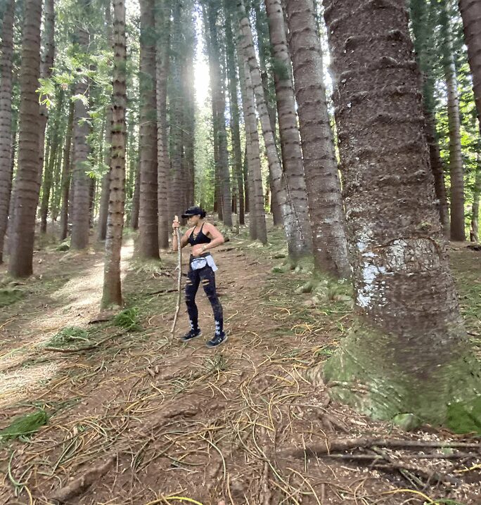 Person standing in a forest with tall trees, sunlight filtering through the leaves, wearing dark clothing and a hat.