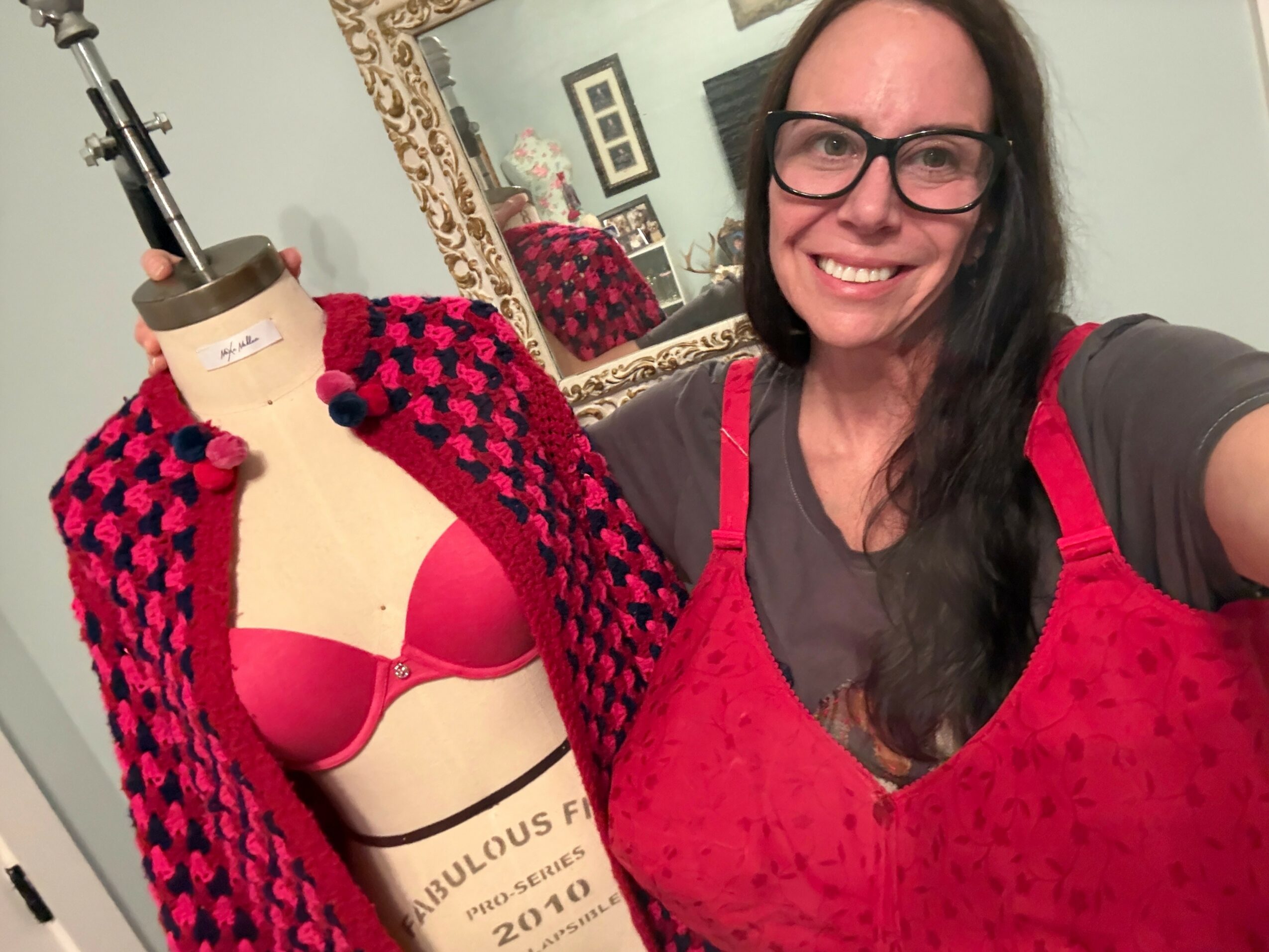 Woman with long dark hair and glasses smiling, wearing a red top, taking a selfie next to a mannequin with a red bra and a pink and black checkered cardigan, in a room with a mirror and framed pictures.