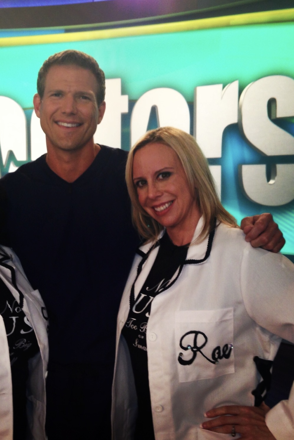 Two smiling people, a man and a woman, standing close together in front of a colorful background, woman wearing a white jacket.