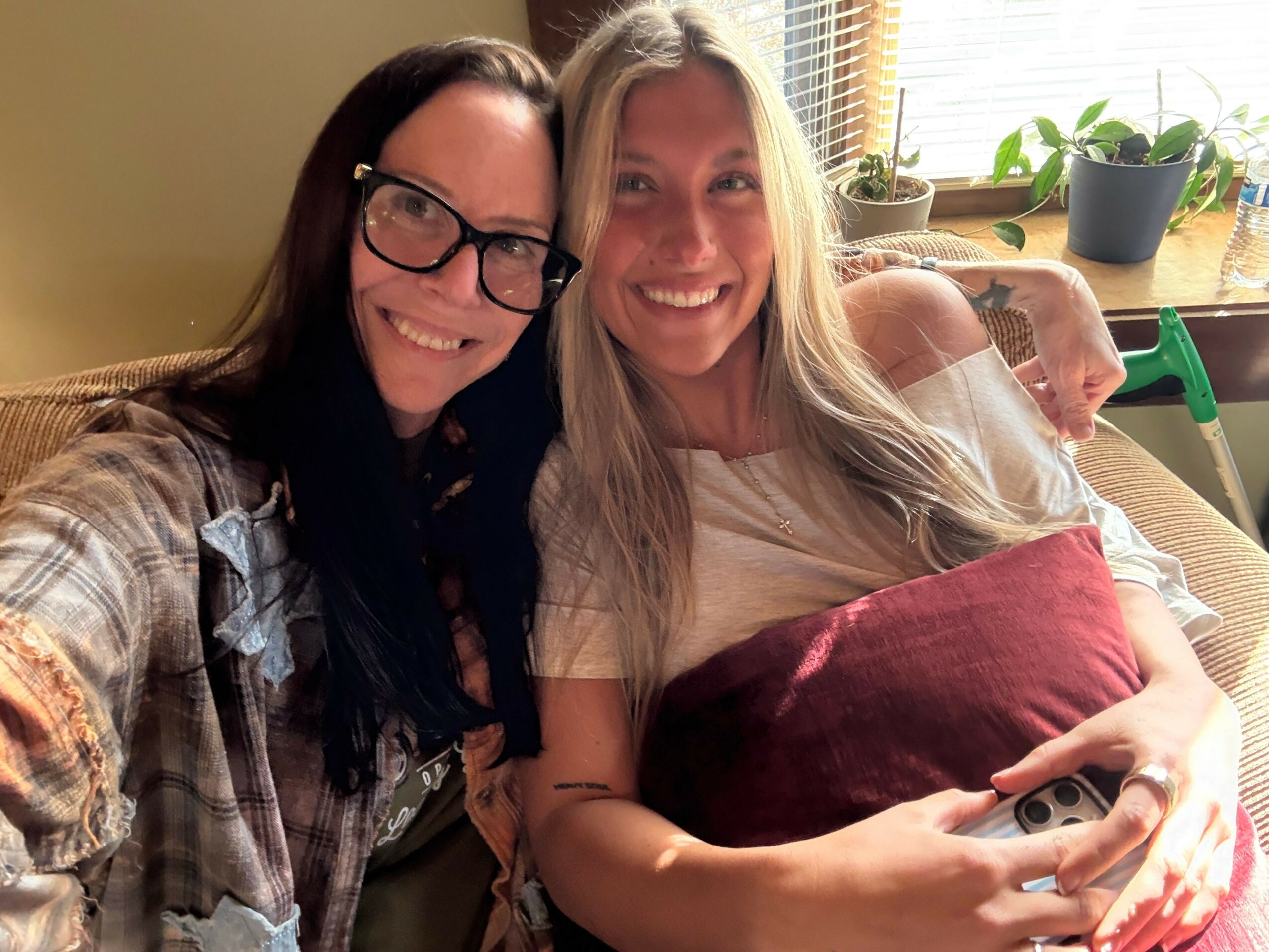 Two women smiling, one with glasses, taking a selfie indoors with plants and furniture visible in the background.