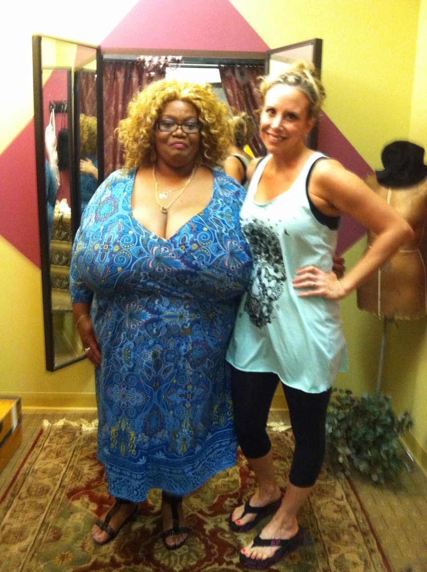 Two women standing in front of a mirror, one wearing a blue patterned dress and glasses, the other in a light blue top and black leggings, smiling.