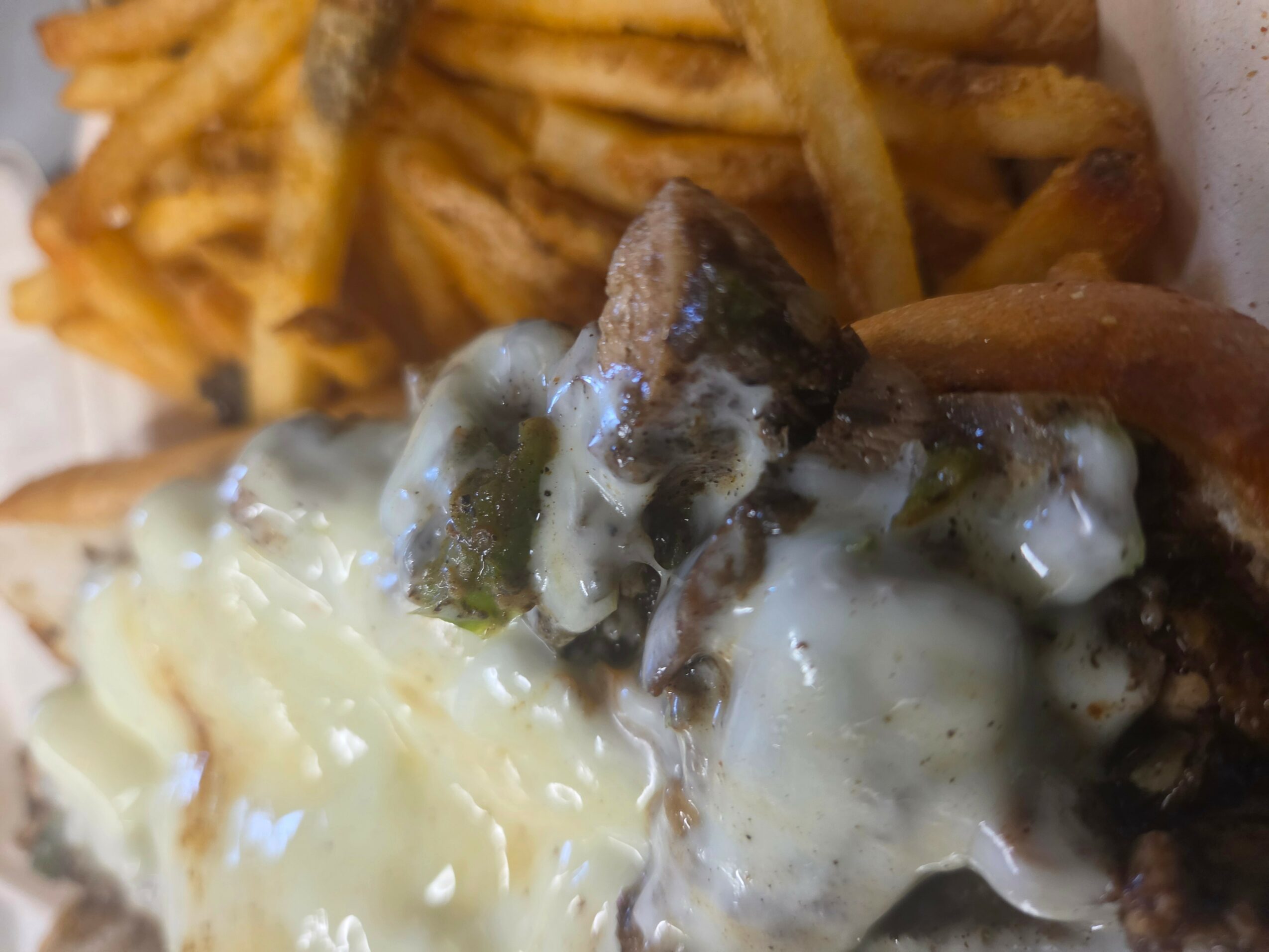 Close-up of a burger with melted cheese, meat, and toppings, with French fries in the background.