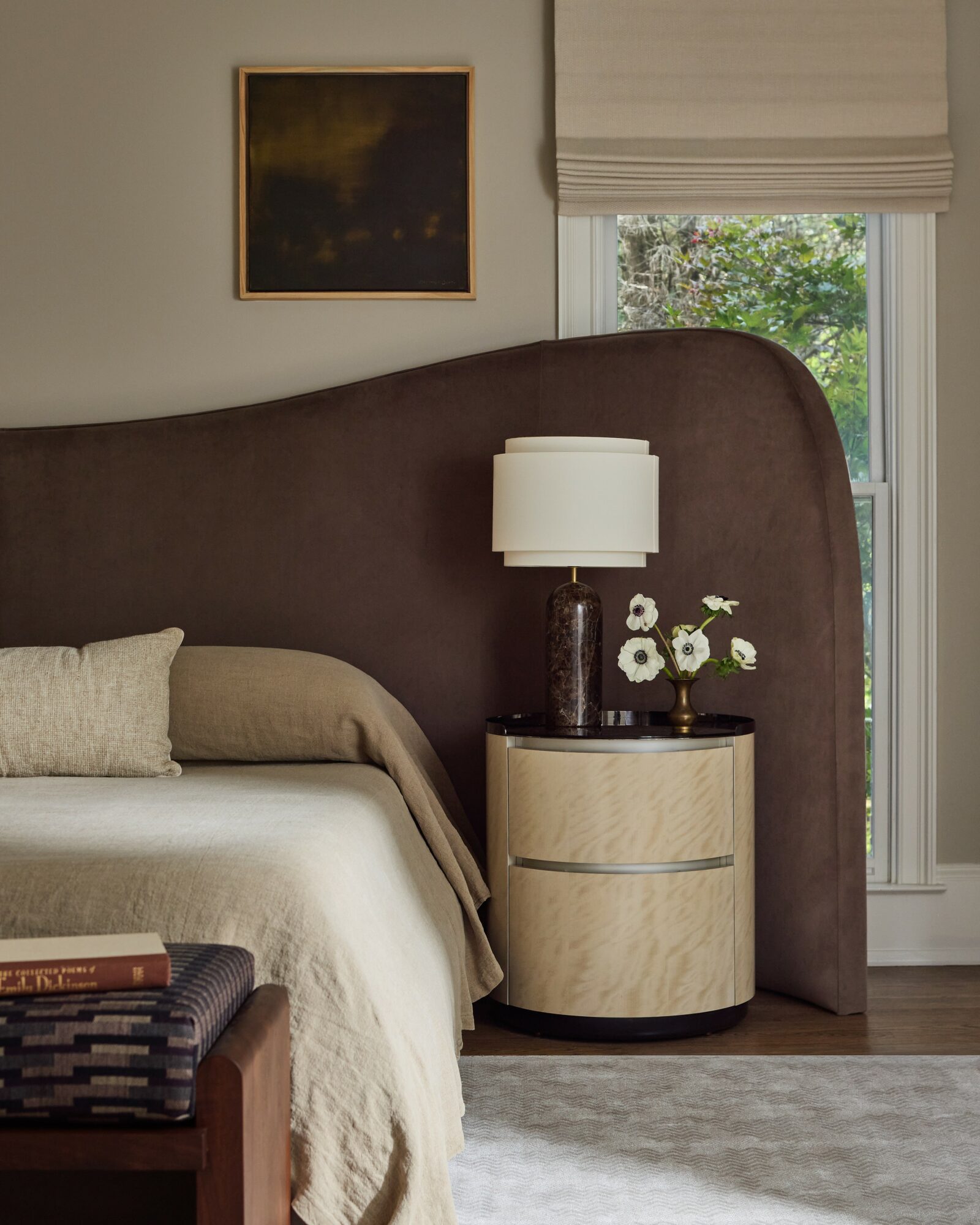Bedroom with a curved headboard, nightstand with a lamp and flowers, window with blinds, and a framed picture on the wall.