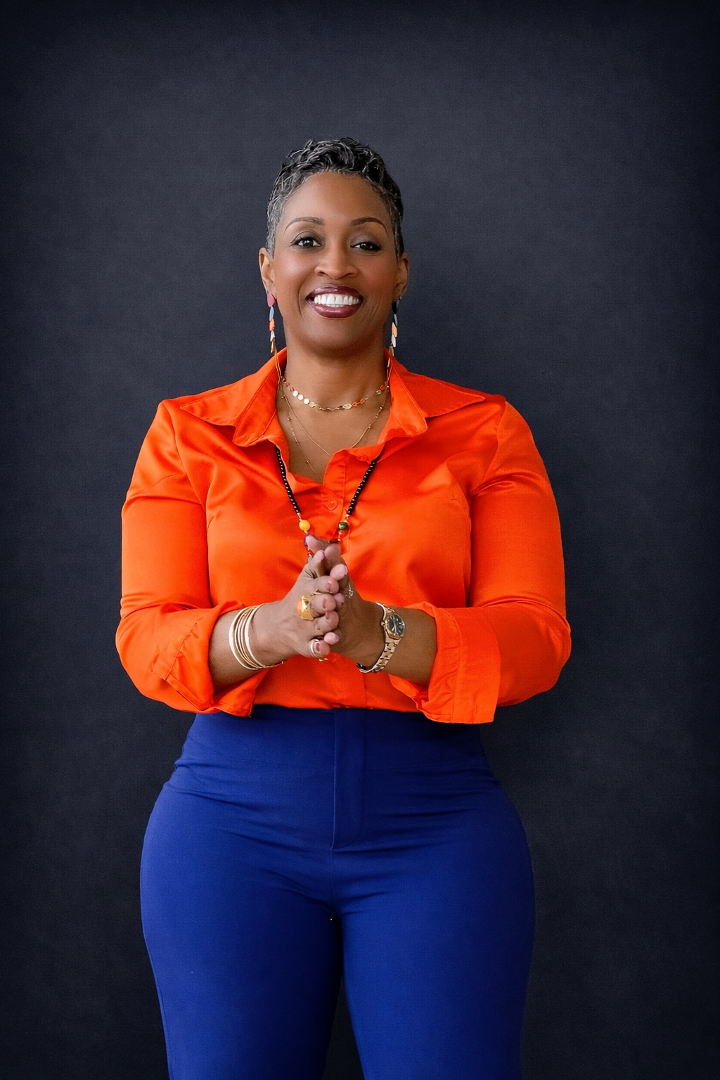 Woman in orange blouse and blue pants smiling with hands clasped, dark background.