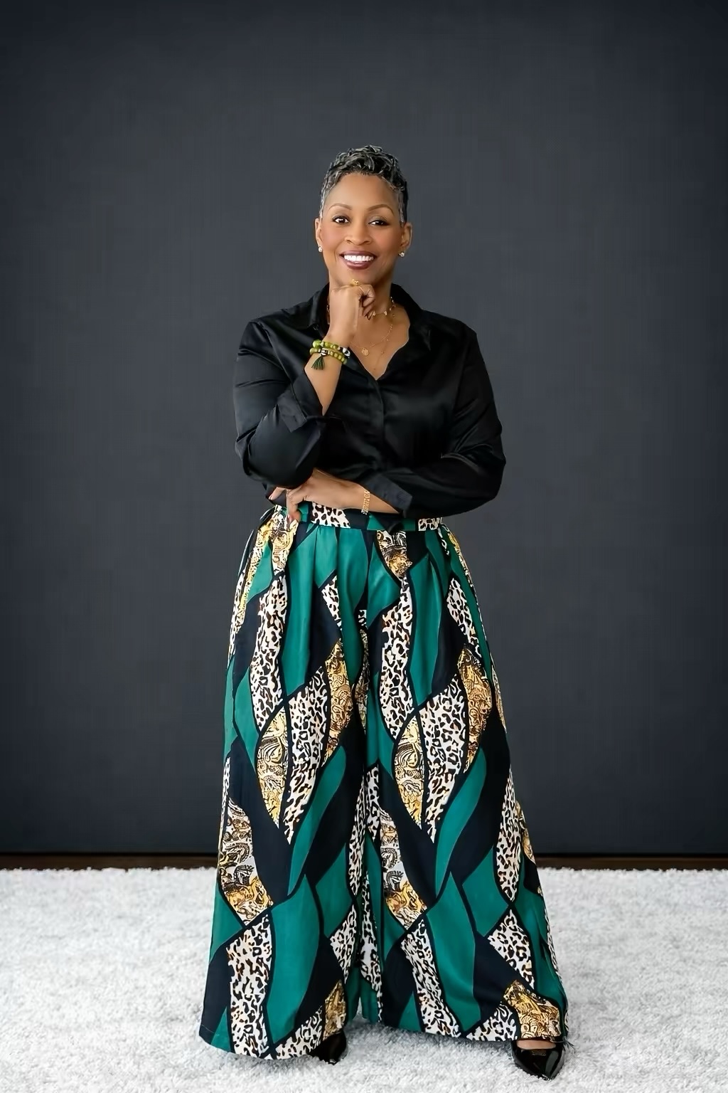 Woman standing with arms crossed, smiling, wearing a black top and patterned wide-leg pants, against a dark background.