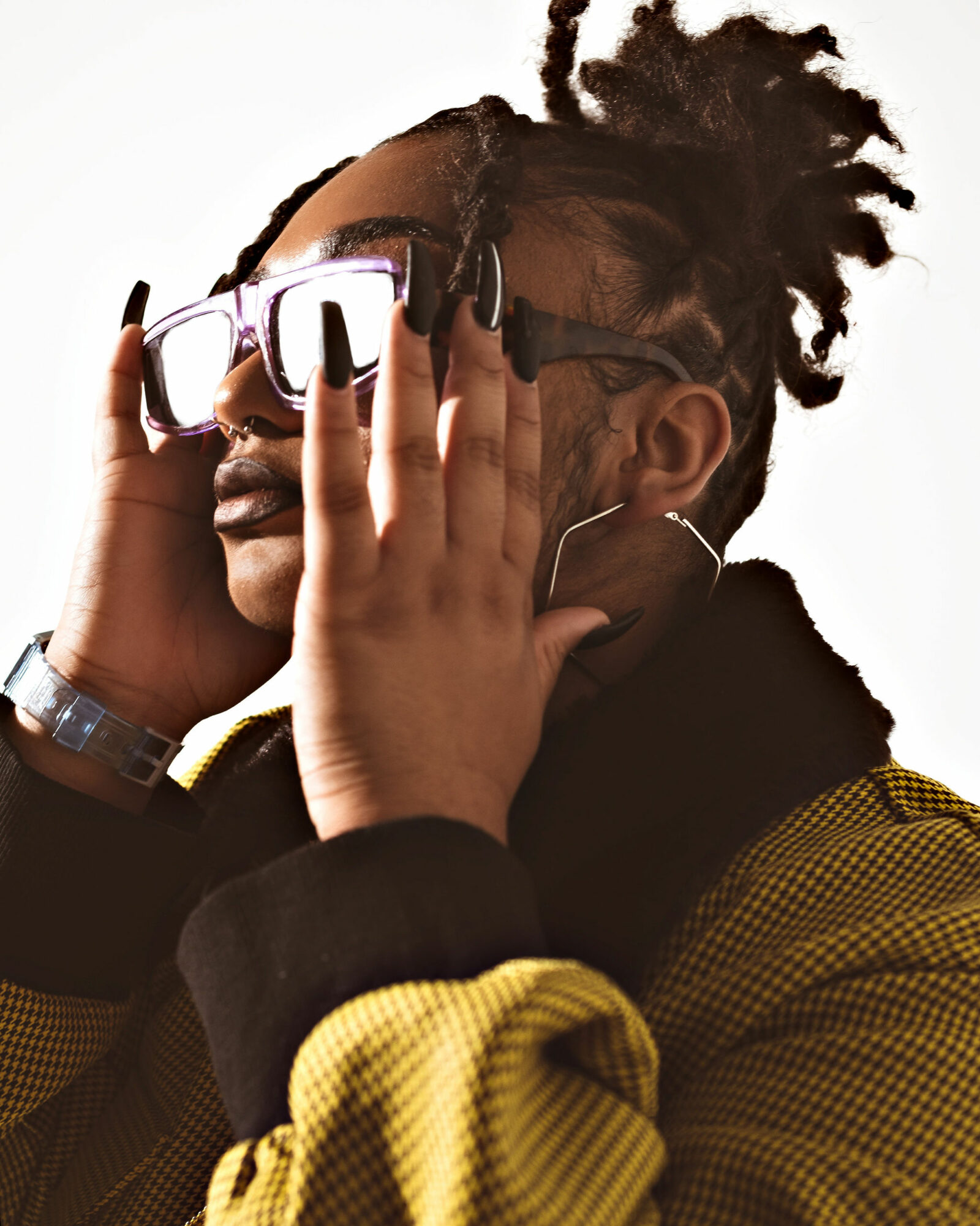 Person with glasses and dreadlocks adjusting glasses, wearing a yellow and black jacket, against a white background.