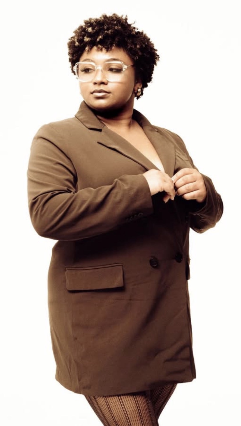 Woman with curly hair and glasses wearing a brown blazer, standing with hands near her chest, looking to the side.