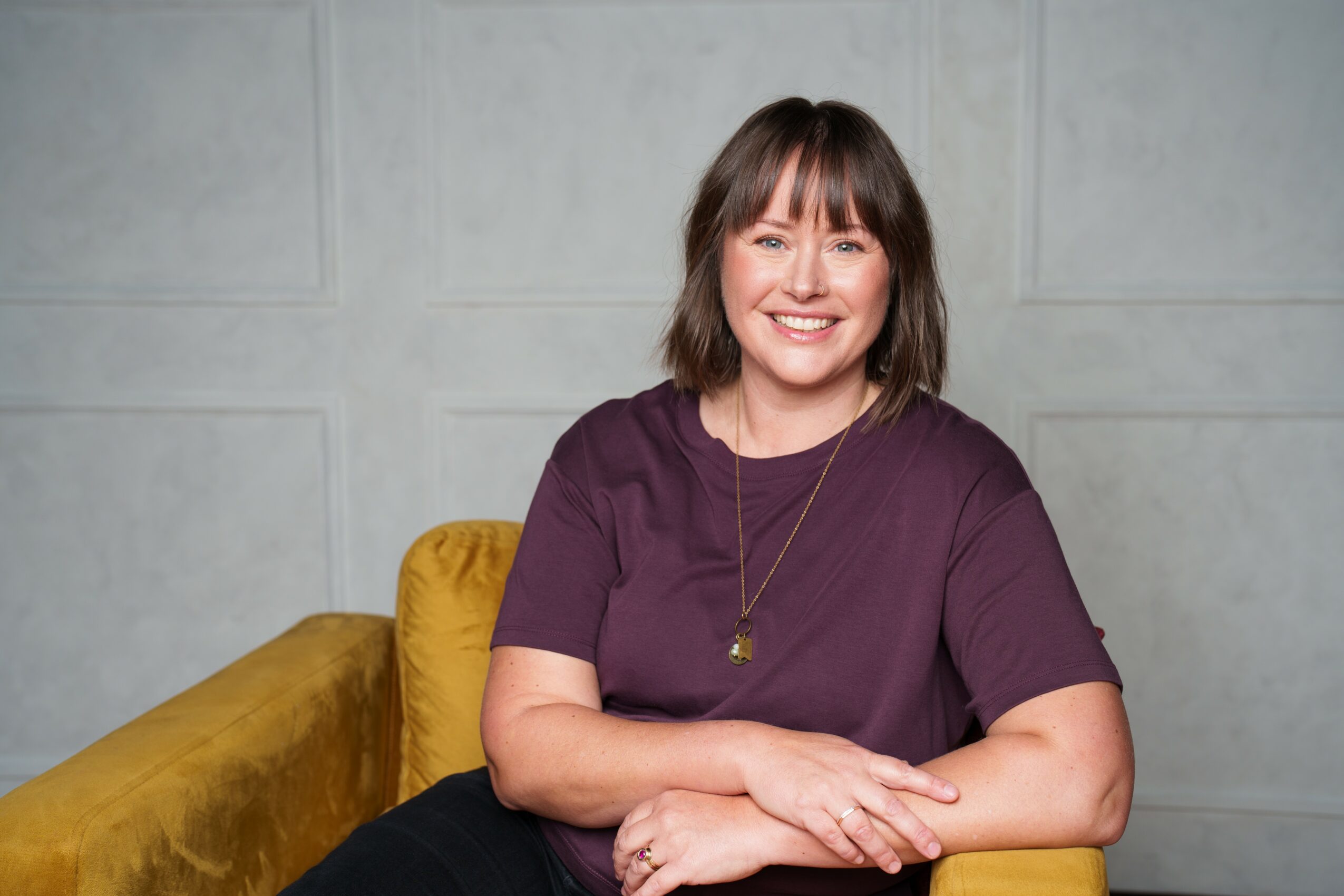 Woman with shoulder-length brown hair and bangs, smiling, wearing a purple top, sitting on a yellow couch against a gray wall.