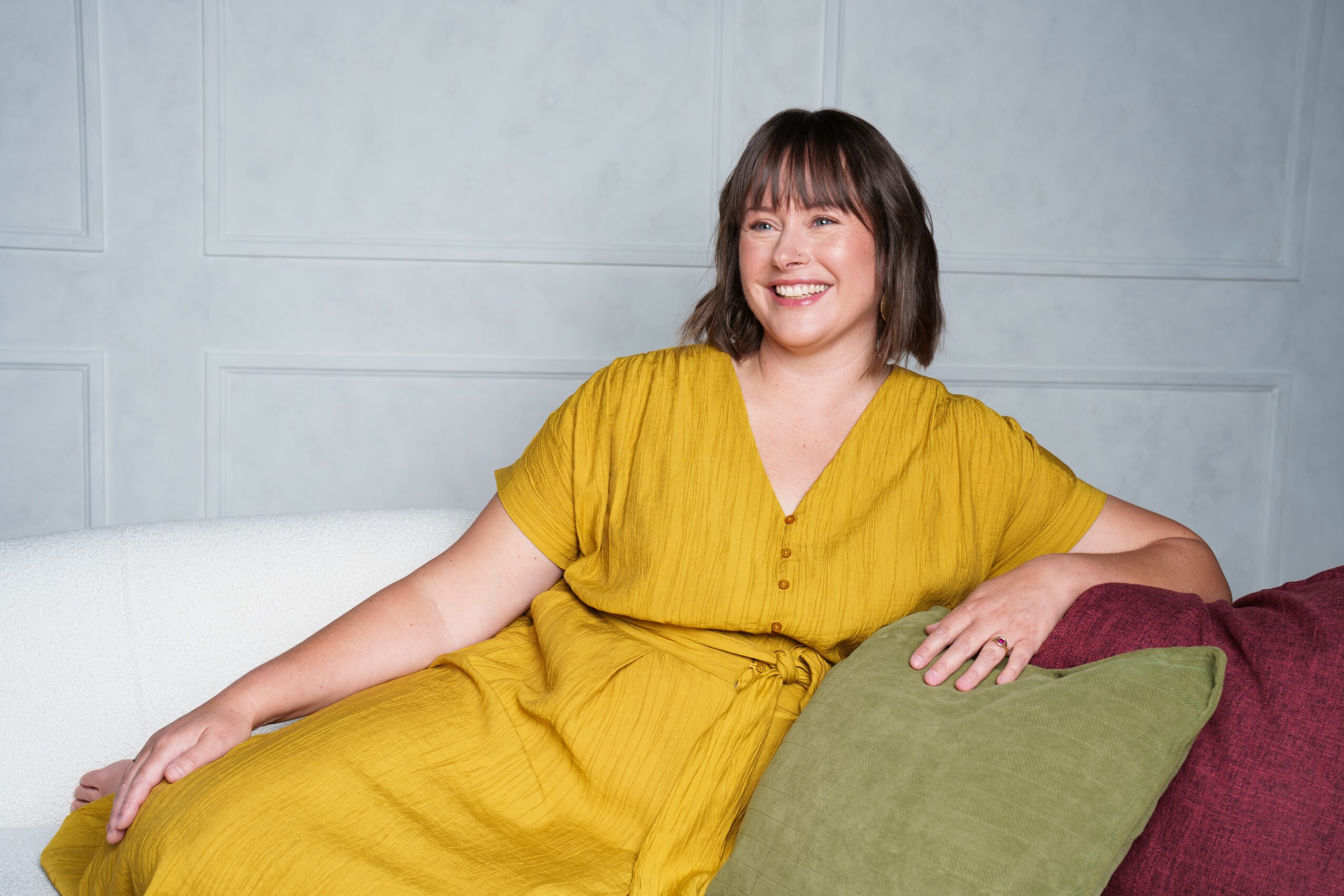 Woman with shoulder-length dark hair smiling, wearing a yellow dress, sitting on a white couch with pillows.