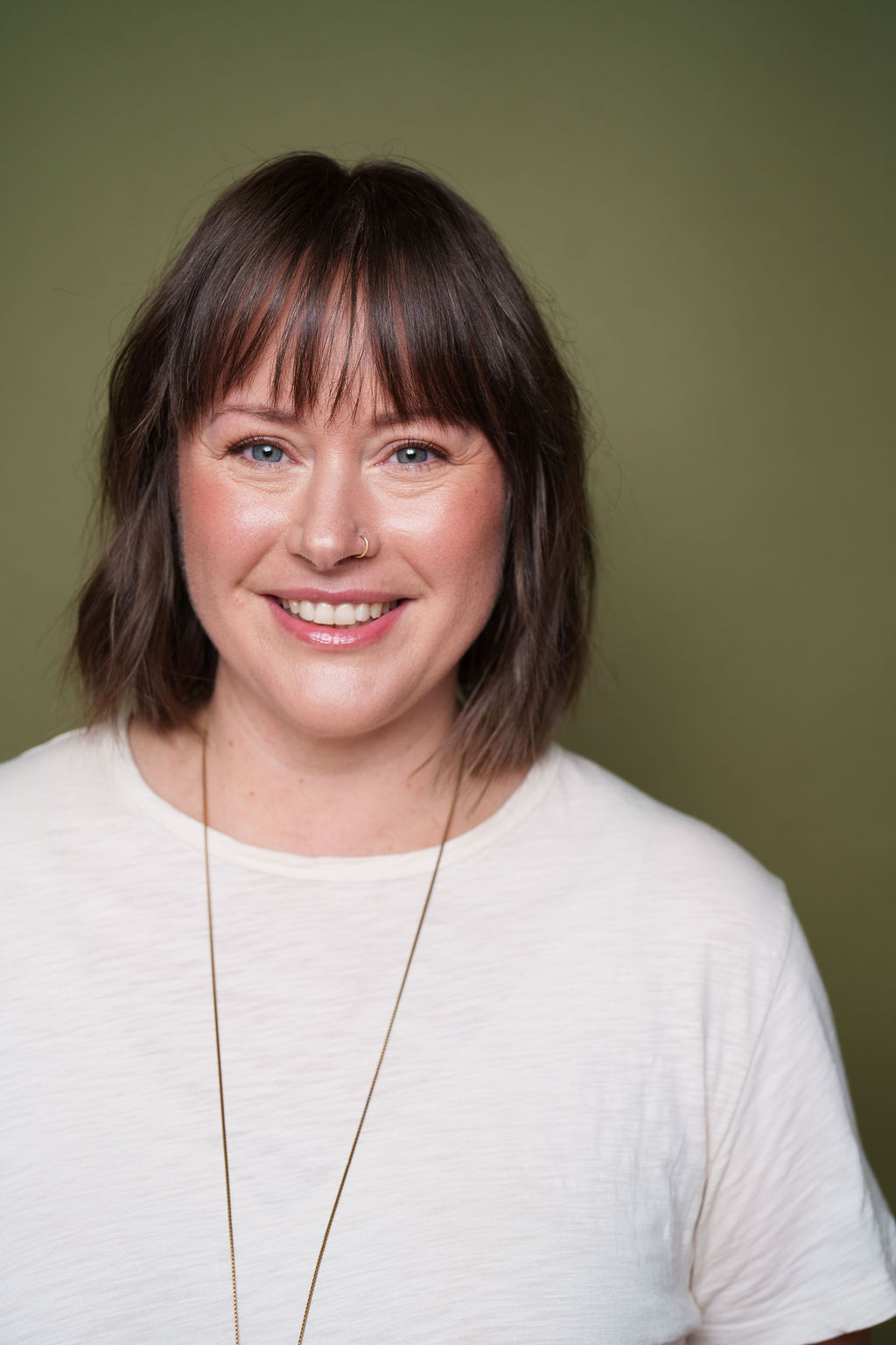 Smiling woman with shoulder-length brown hair and bangs, wearing a white shirt, against a green background.