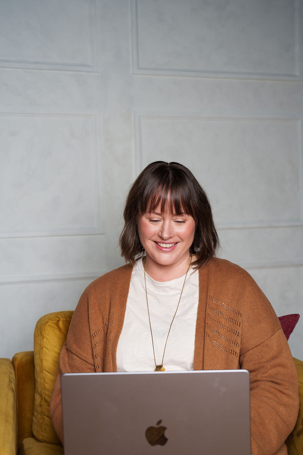 Woman with shoulder-length hair smiling while using a laptop, sitting on a yellow couch against a plain wall.