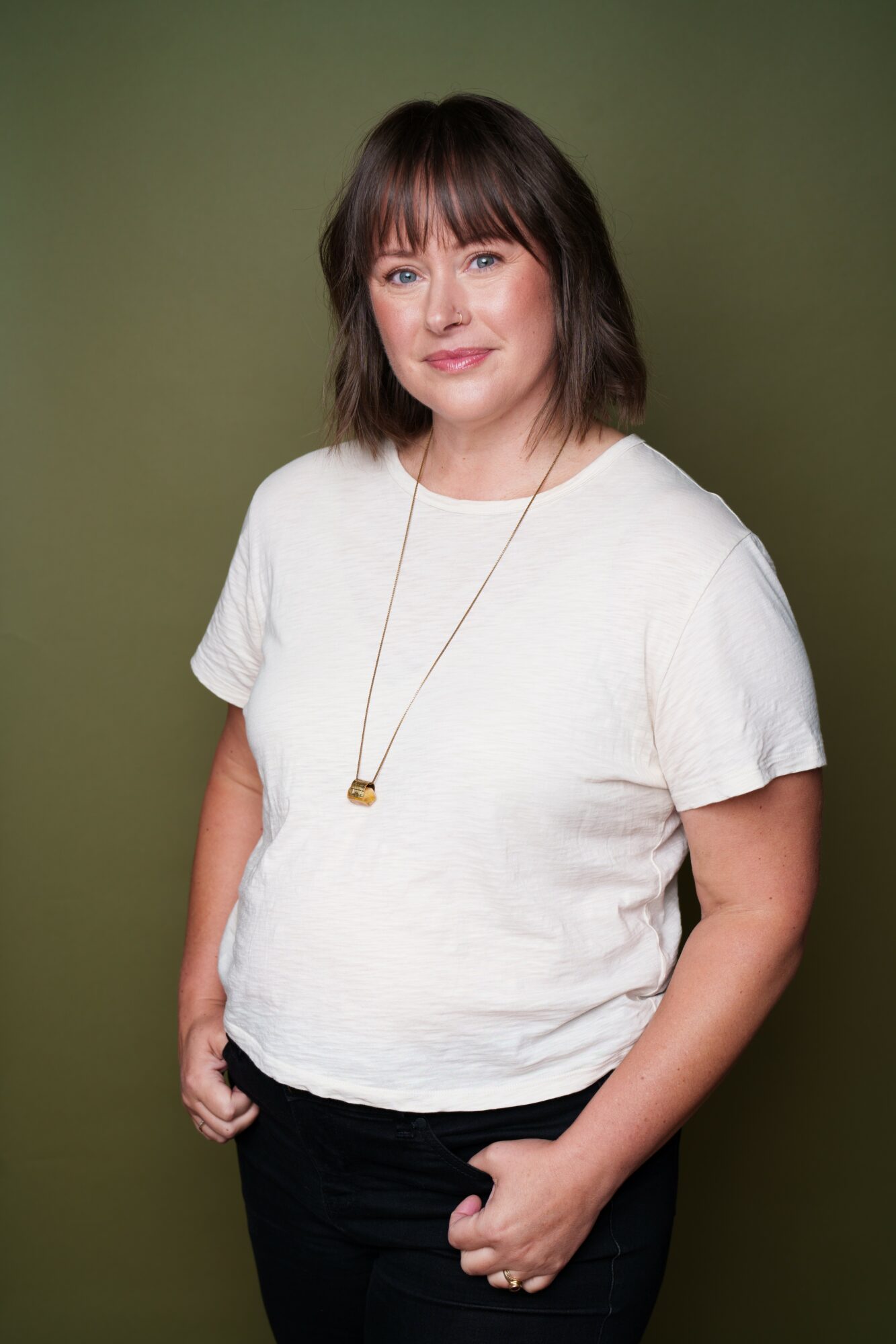 Woman with shoulder-length brown hair and bangs, wearing a white t-shirt and black pants, standing against a green background.