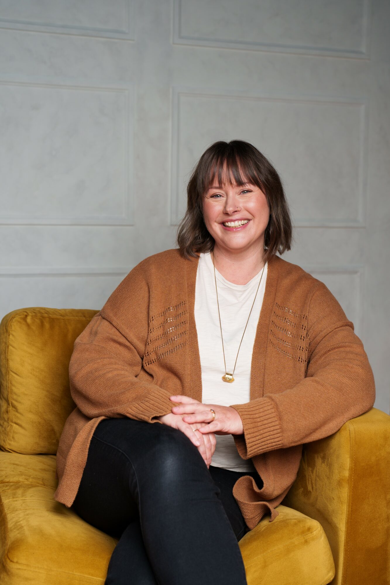 Woman with shoulder-length hair smiling, sitting on a yellow couch, wearing a brown cardigan and white top.