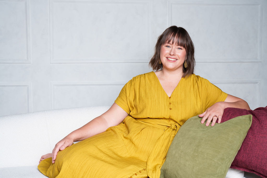 Woman with short hair in a yellow dress sitting on a white couch, smiling, with green and purple pillows, gray wall background.