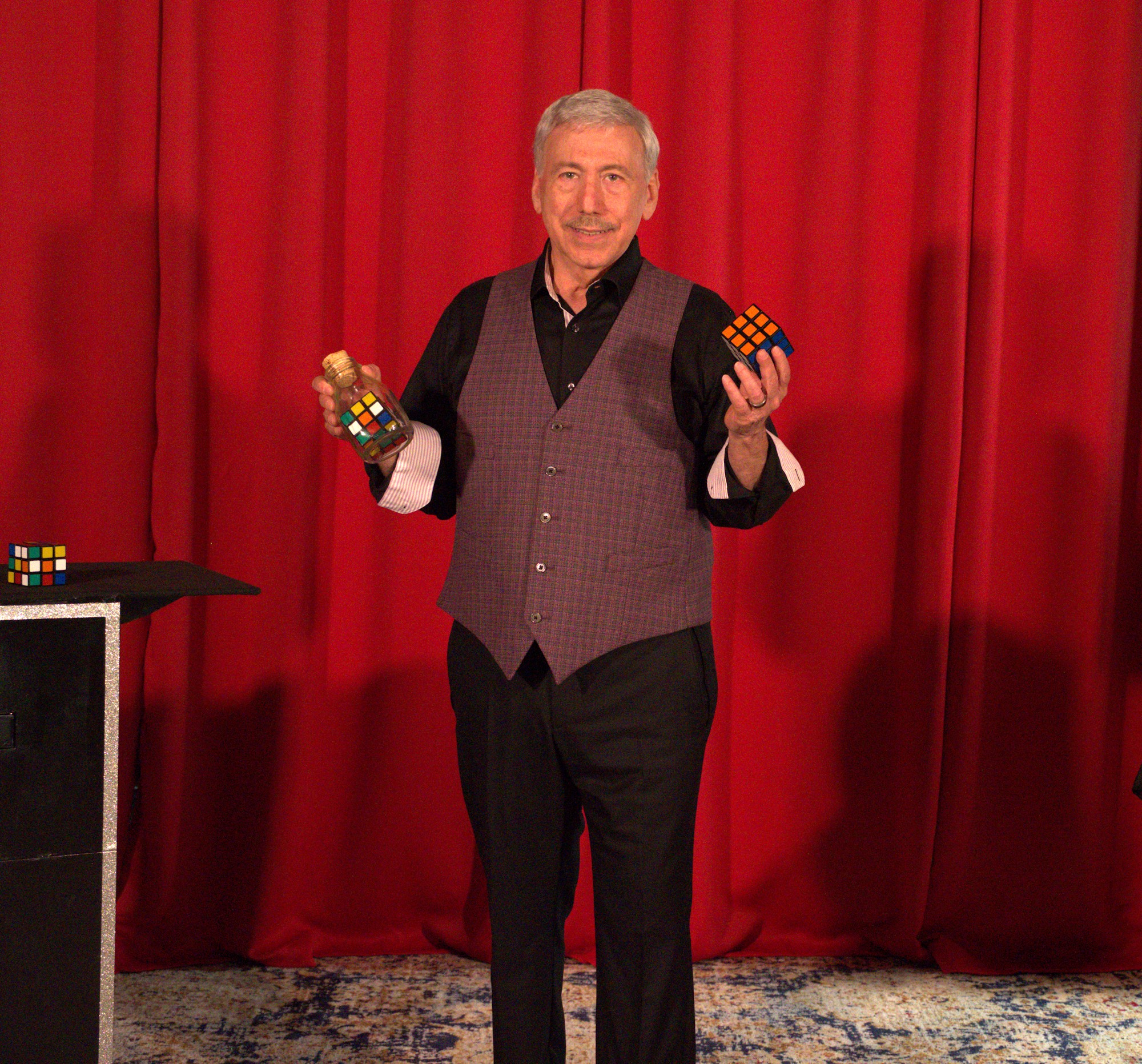 Older man holding two Rubik's cubes in front of red curtains, smiling, wearing a vest and black pants.