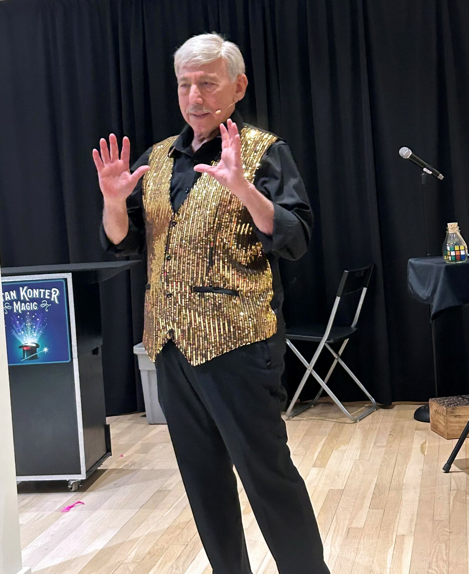 Older man with white hair wearing a gold sequin vest and black outfit, gesturing with hands in a room with black curtains.