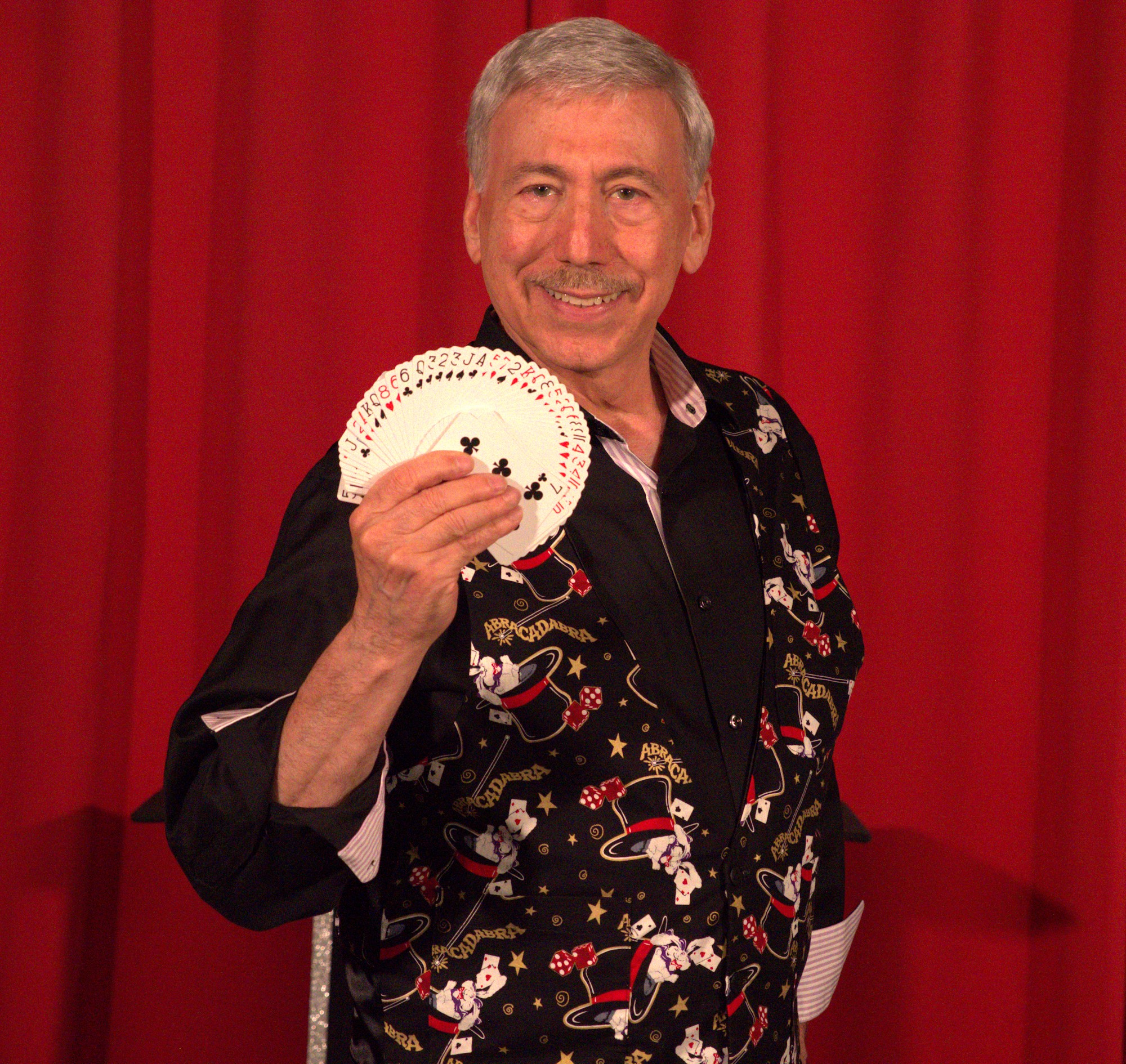 Older man smiling, holding a fan, standing in front of red curtains, wearing a festive black shirt with holiday patterns.