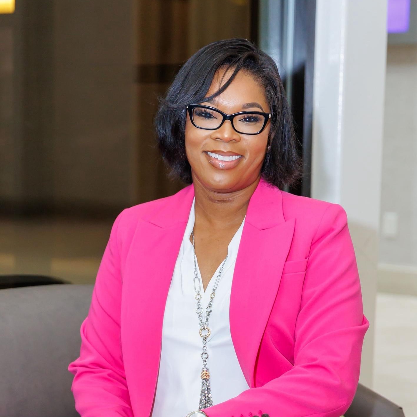 Woman with glasses smiling, wearing a pink blazer and white blouse, indoors with a neutral background.
