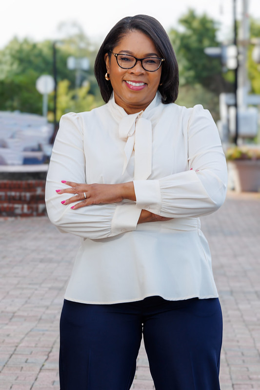 Woman with glasses and earrings smiling, arms crossed, outdoors in a city setting, wearing a white blouse and dark pants.