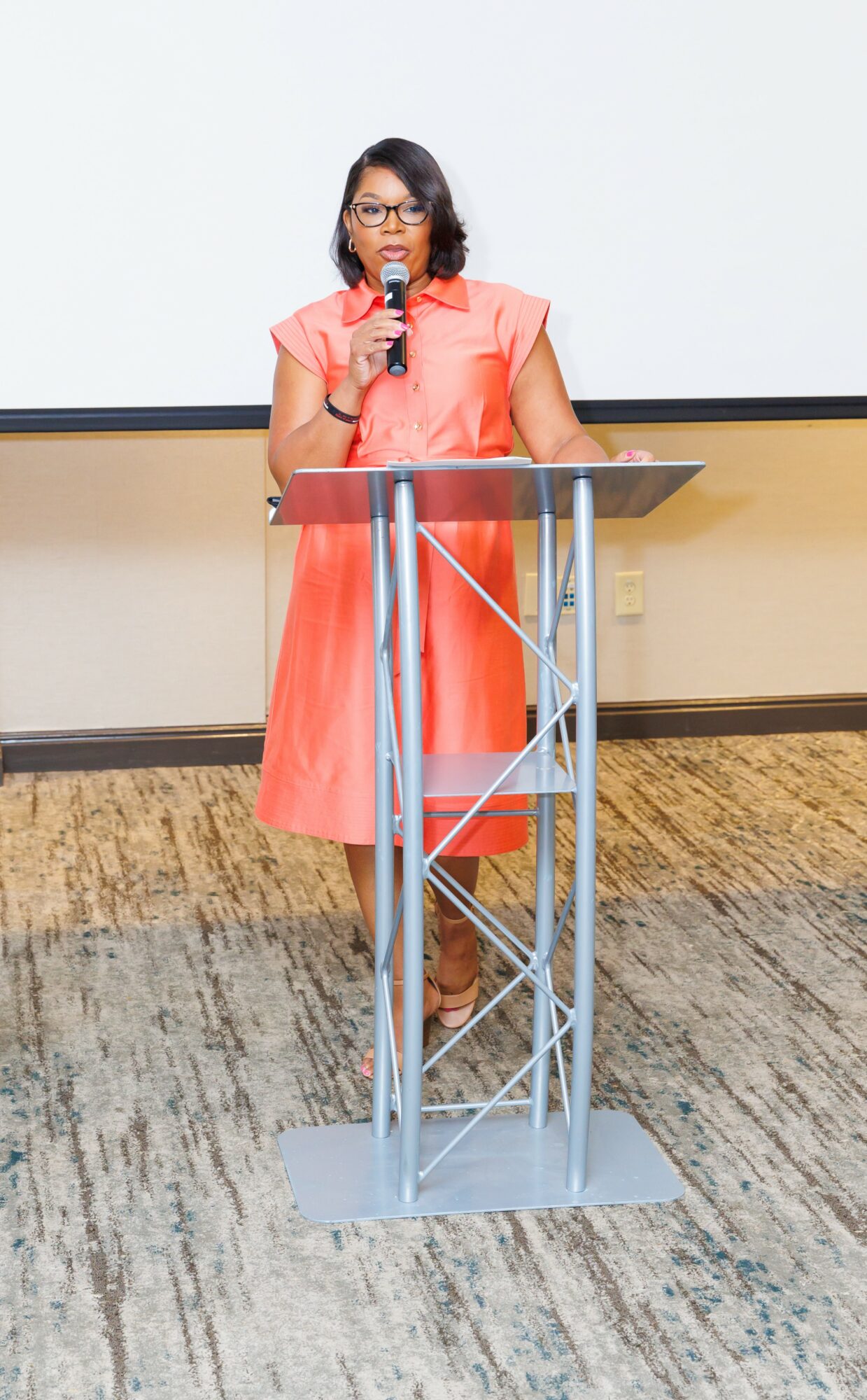 Woman in coral dress speaking into a microphone at a podium in a room with a white wall and carpeted floor.