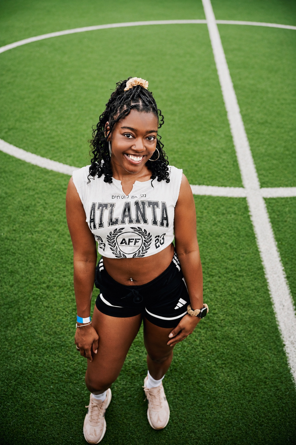 Young woman standing on a green sports field, smiling, wearing athletic clothing and sneakers.