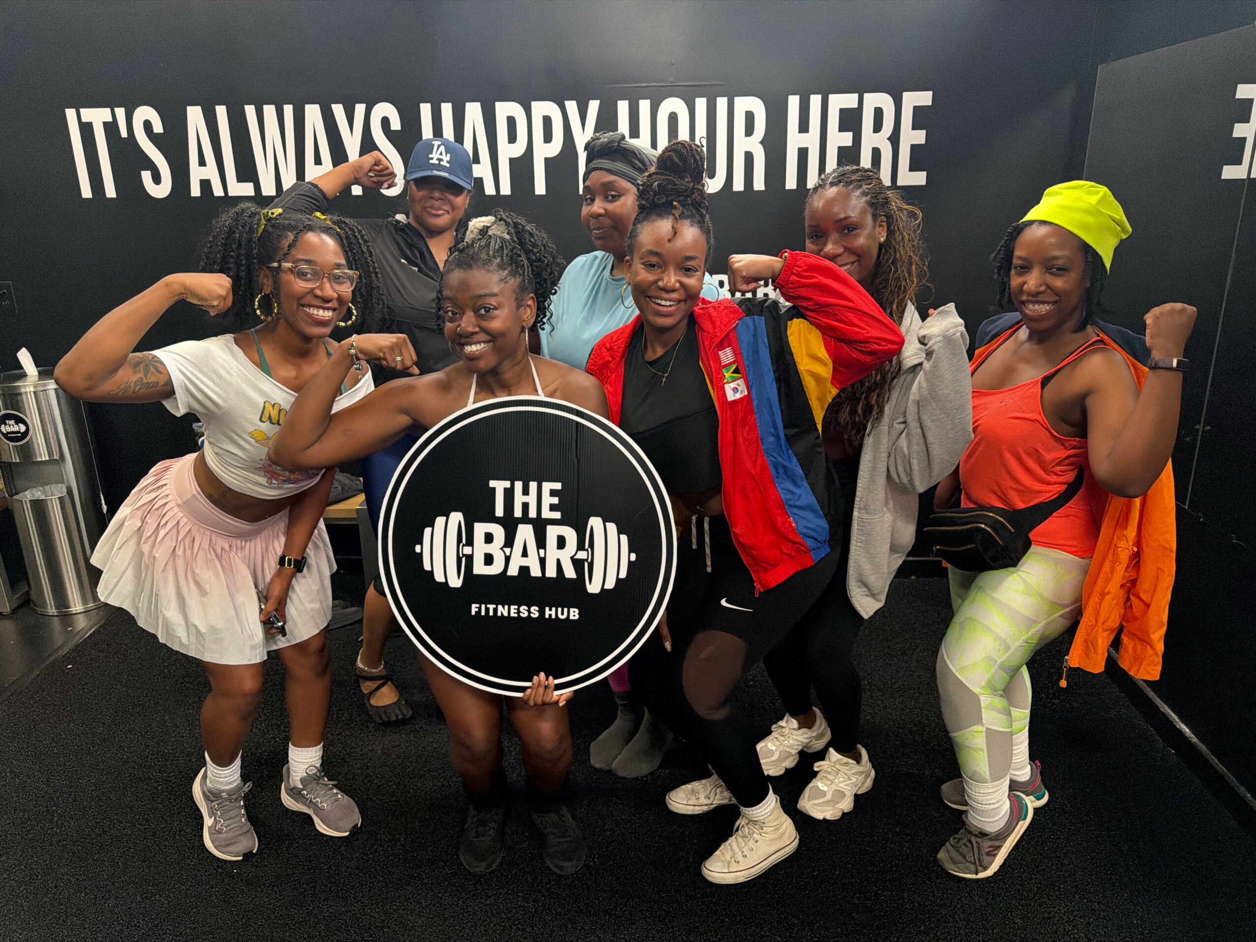 Group of seven smiling women and girls posing indoors, holding a circular sign that reads 'The Bar Fitness Hub'.