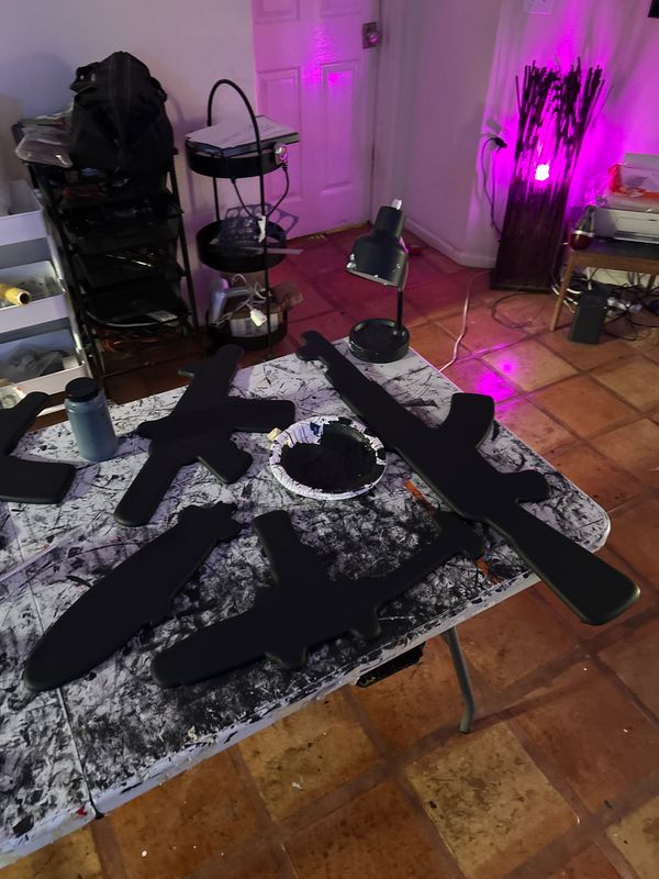 Table with black firearms and accessories, room with purple lighting, curtains, and various objects in the background.