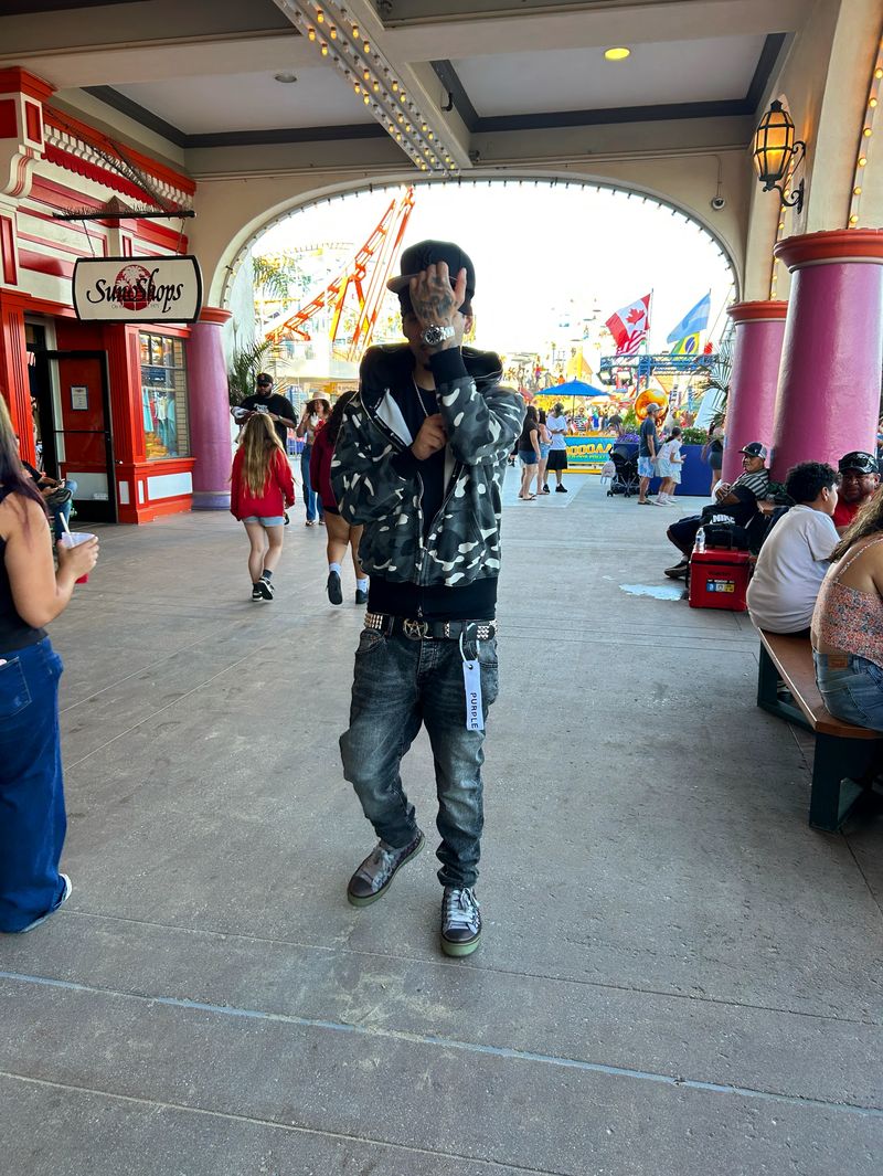 Person standing in front of an amusement park entrance, holding a phone, with rides and visitors in background.